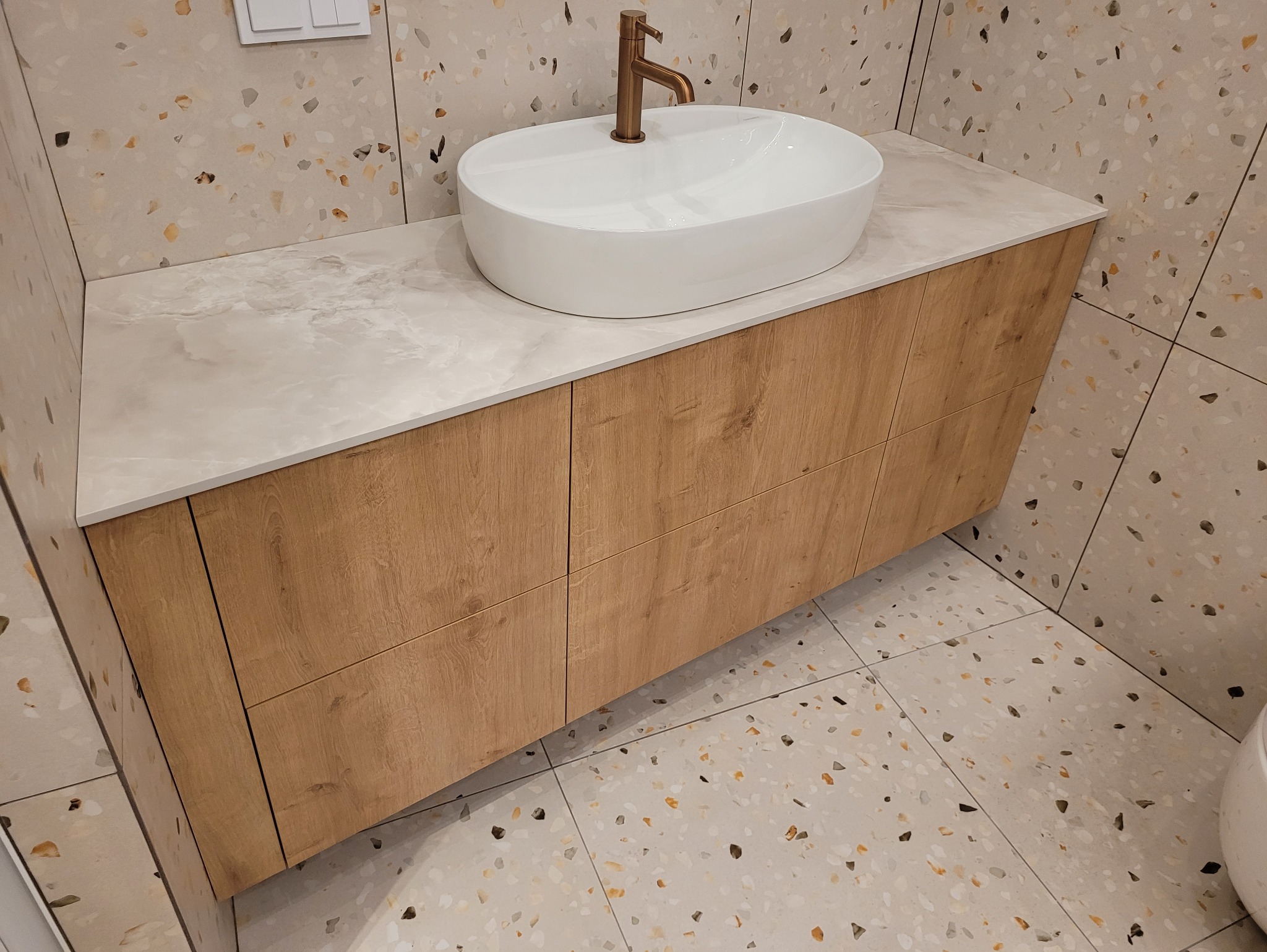 Nowoczesna szafka łazienkowa z drewnianymi frontami i blatem imitującym marmur, na którym stoi owalna umywalka i bateria w kolorze miedzi. Tło stanowią płytki lastryko.