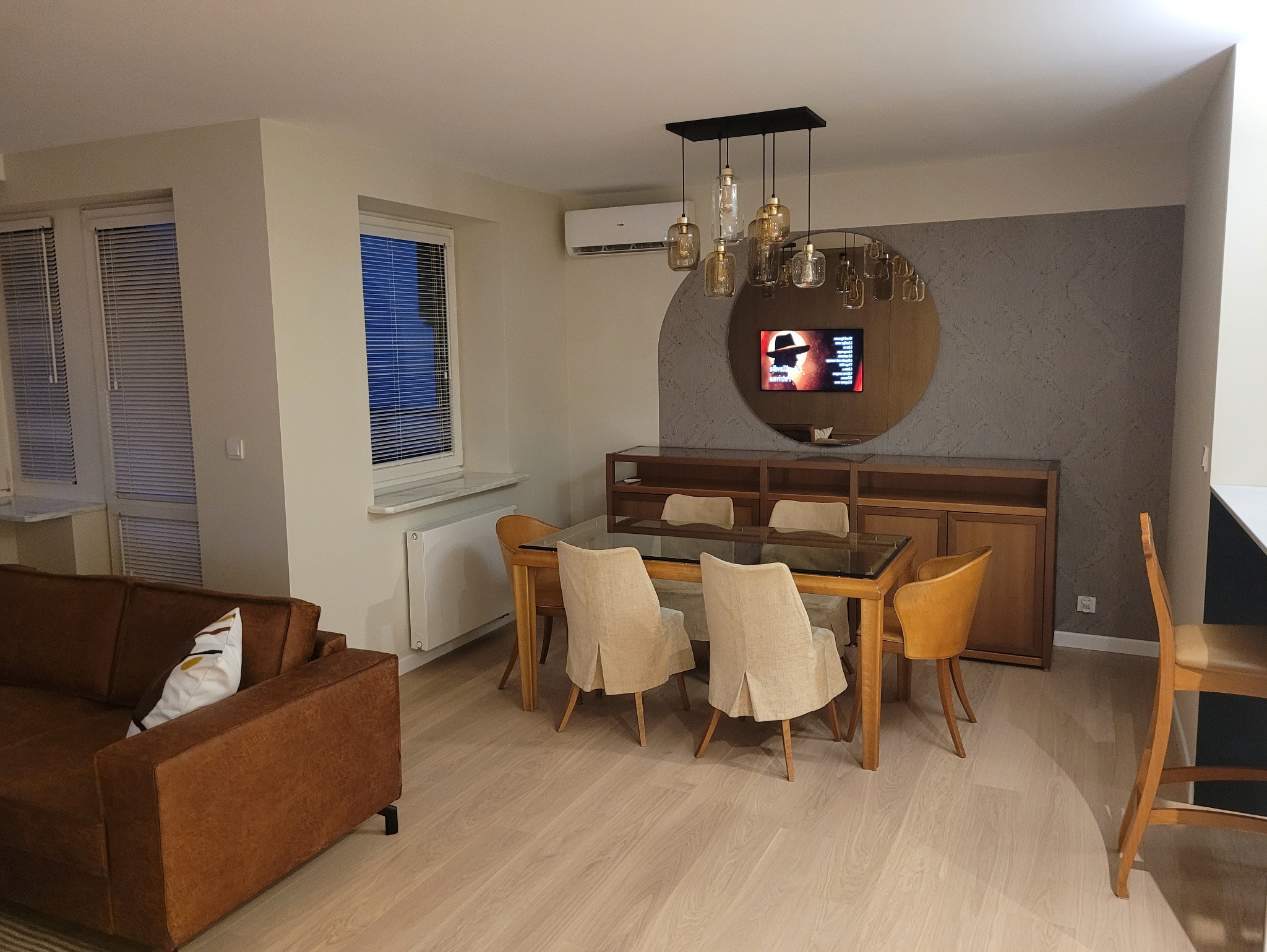 Jasne wnętrze jadalni z drewnianym stołem i krzesłami, nad którymi wisi designerska lampa. W tle szafka i okrągłe lustro z telewizorem.