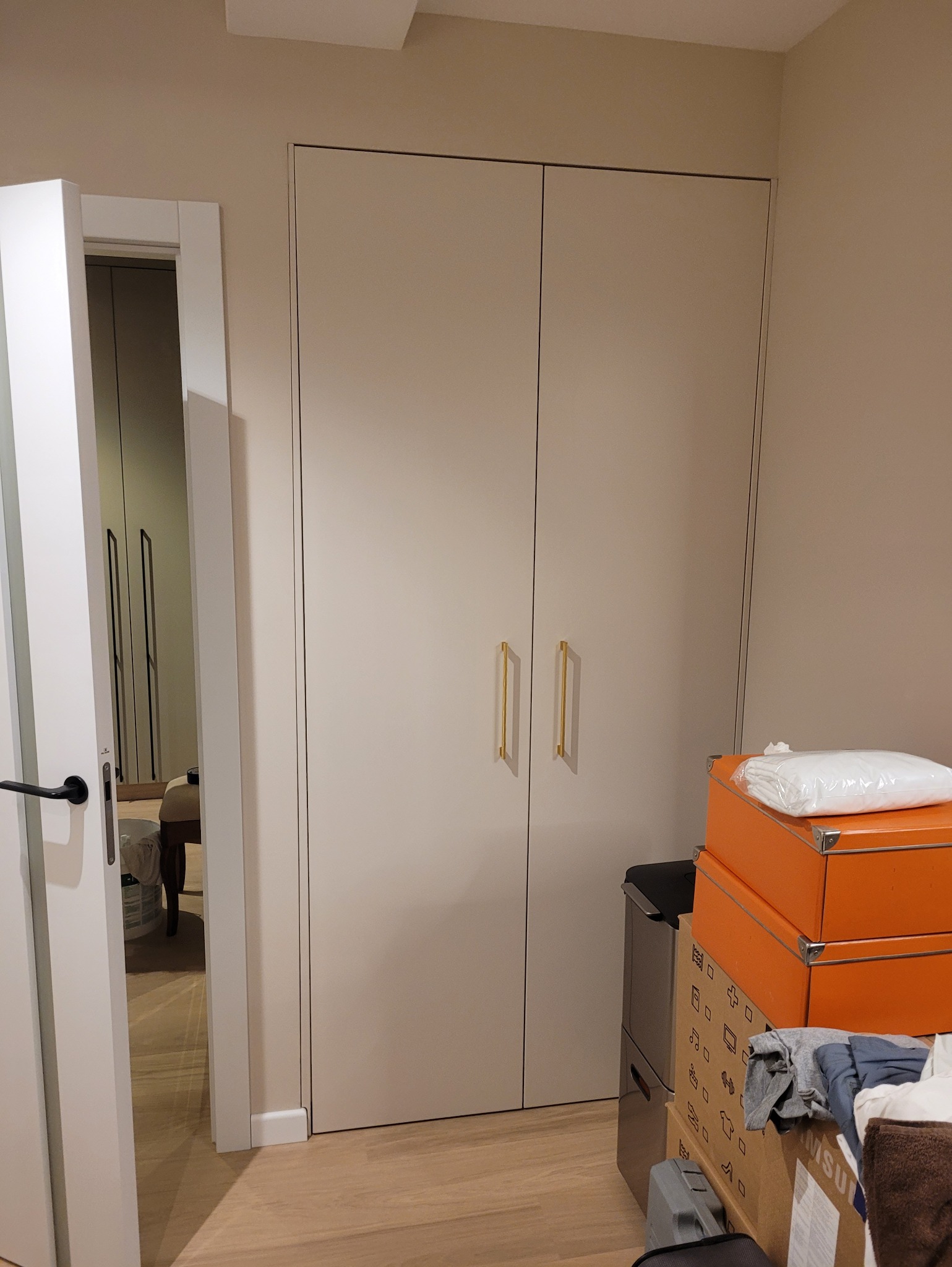 Biała szafa wnękowa z złotymi uchwytami, widoczna z boku, z otwartymi drzwiami po lewej stronie, ukazującymi wnętrze innej szafy. U dołu kartony i sprzęt.