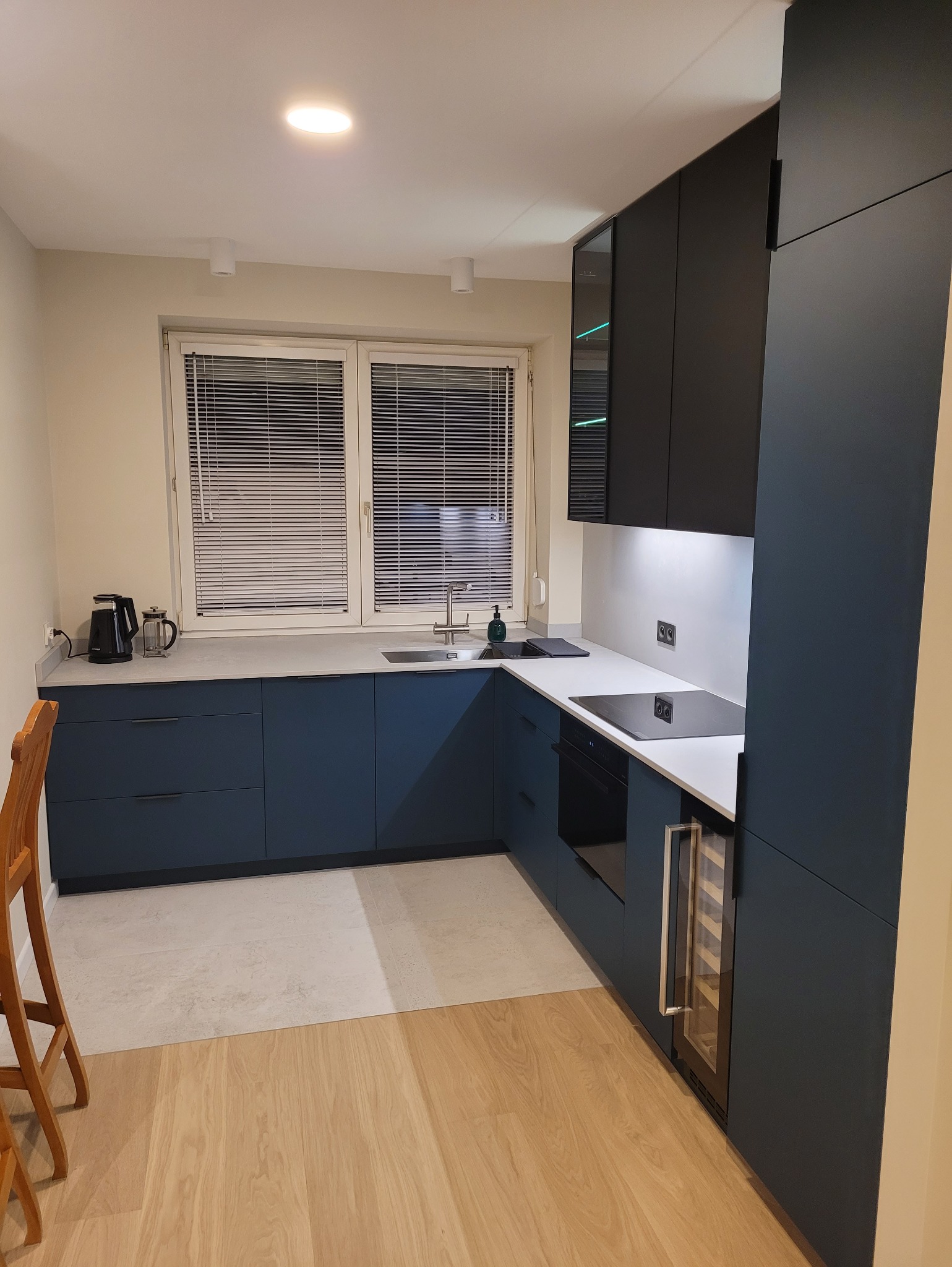 Nowoczesna kuchnia w Osiecku z granatowymi szafkami dolnymi i czarnymi górnymi. Jasny blat i podłoga. Widoczna płyta indukcyjna i lodówka na wino.