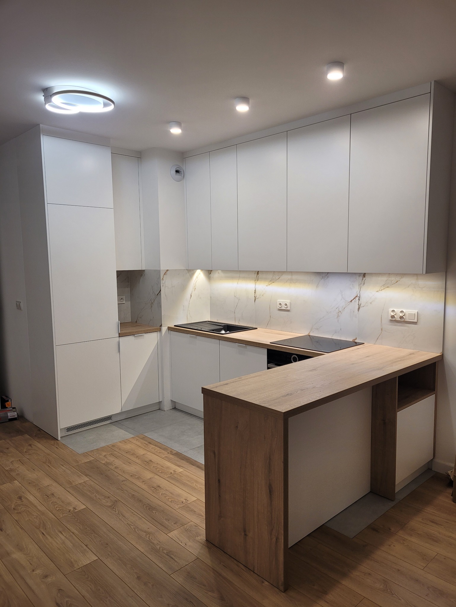 Nowoczesna kuchnia na wymiar z białymi szafkami i drewnianym blatem, oświetlona punktowo, z marmurowym panelem nad blatem i wyspą kuchenną.