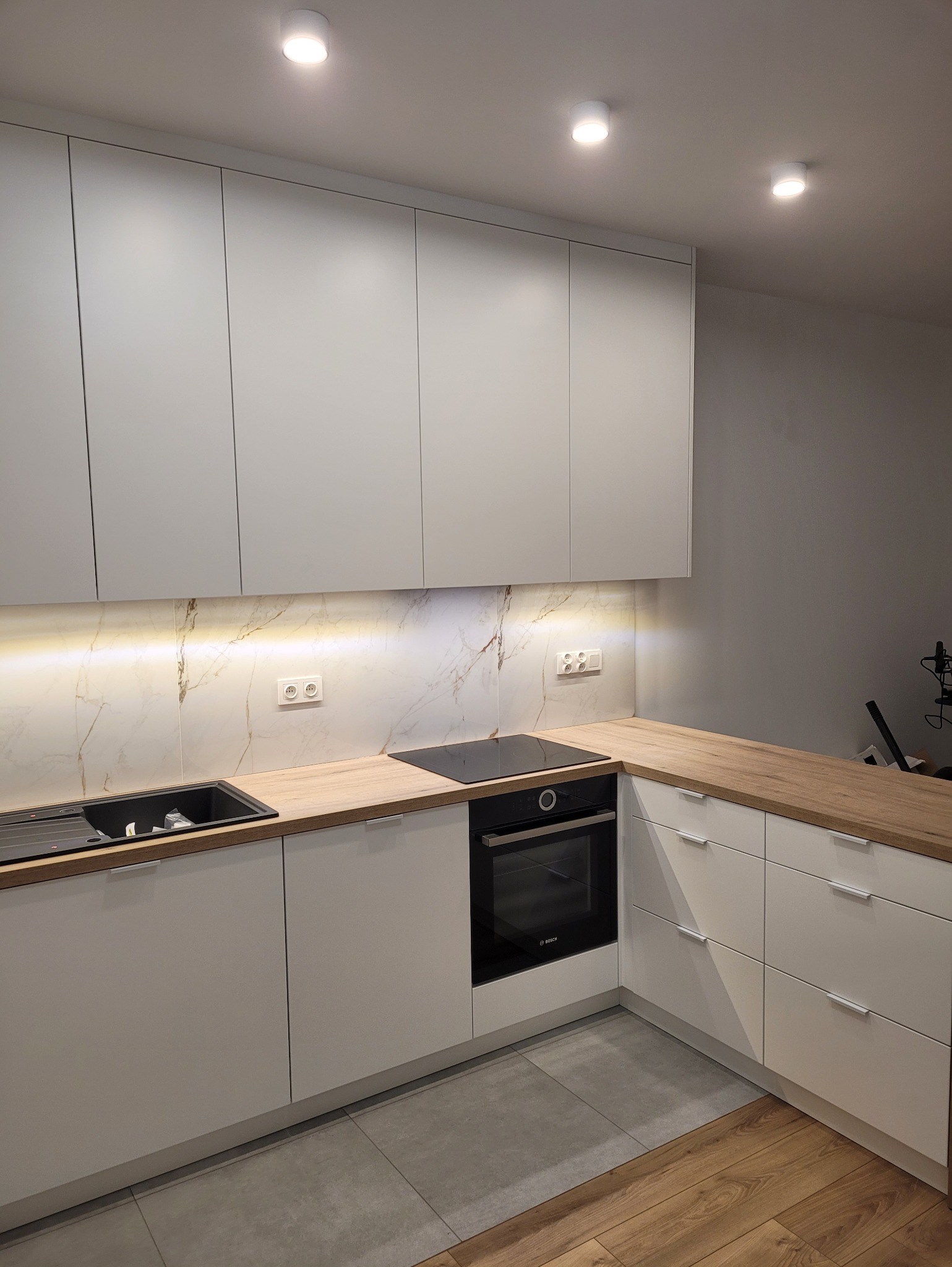 Nowoczesna kuchnia z białymi frontami, drewnianym blatem i marmurowym panelem. Oświetlenie LED podkreśla detale. Widoczny piekarnik Bosch i zlewozmywak.
