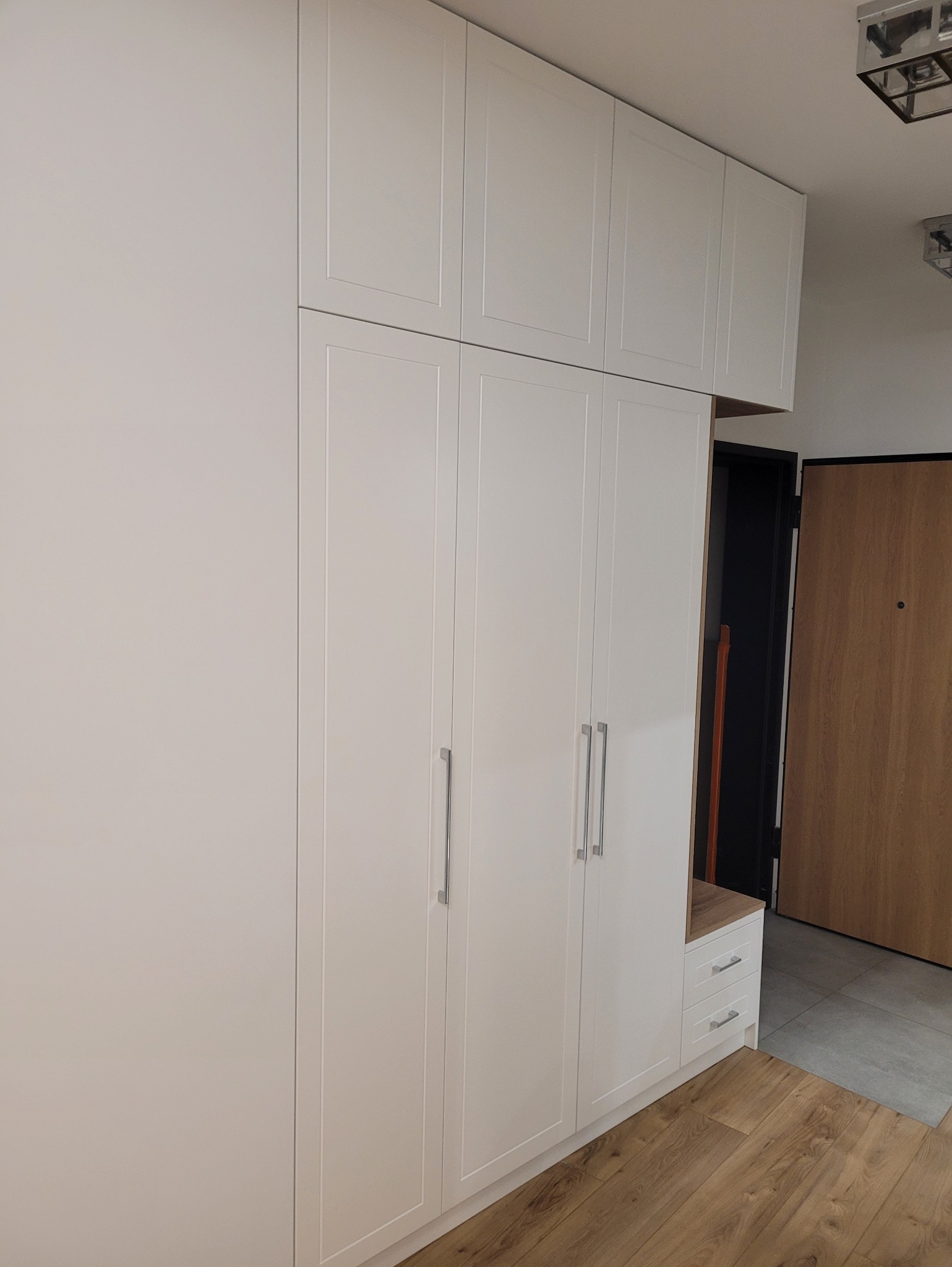 Biała szafa wnękowa z czterema drzwiami i górnymi szafkami, zintegrowana z siedziskiem i szufladami, w jasnym przedpokoju z drewnianą podłogą i drzwiami.