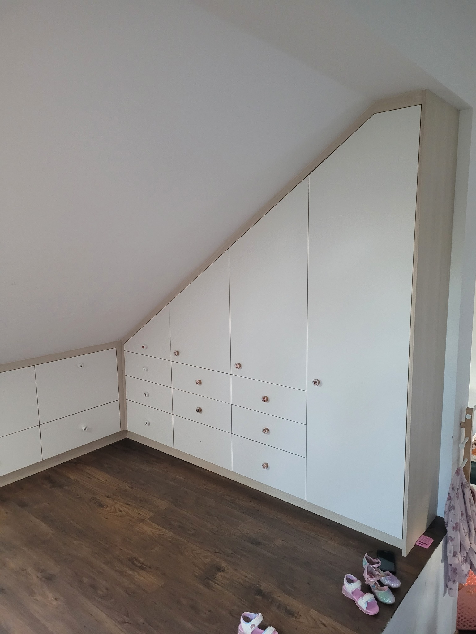 Biała szafa wnękowa na poddaszu ze skośnym sufitem, z drewnianą podłogą i dziecięcymi sandałkami. Szafki z okrągłymi uchwytami.
