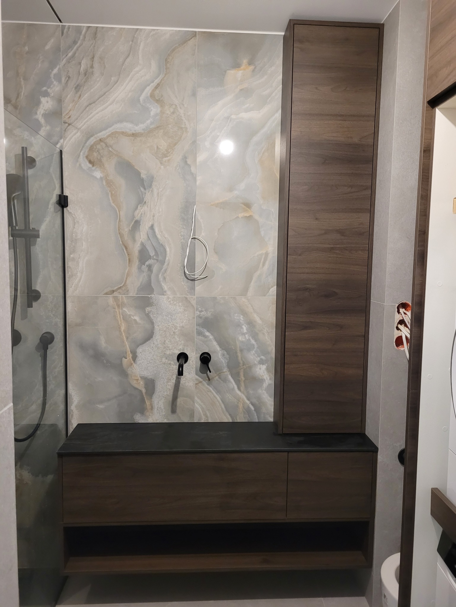 Nowoczesna łazienka z szafką pod umywalkę i wysoką szafką w kolorze ciemnego drewna, na tle marmurowych płytek. Widoczne czarne baterie i elementy prysznica.