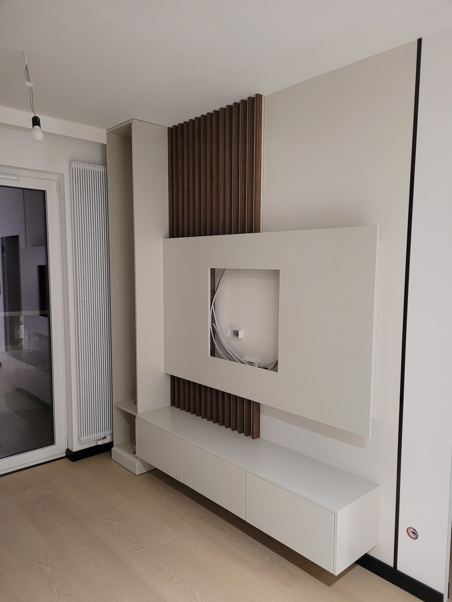 Nowoczesna zabudowa ściany TV z drewnianymi lamelami i białymi szafkami. Minimalistyczny design, widoczne okablowanie w otworze na telewizor. Jasne wnętrze.