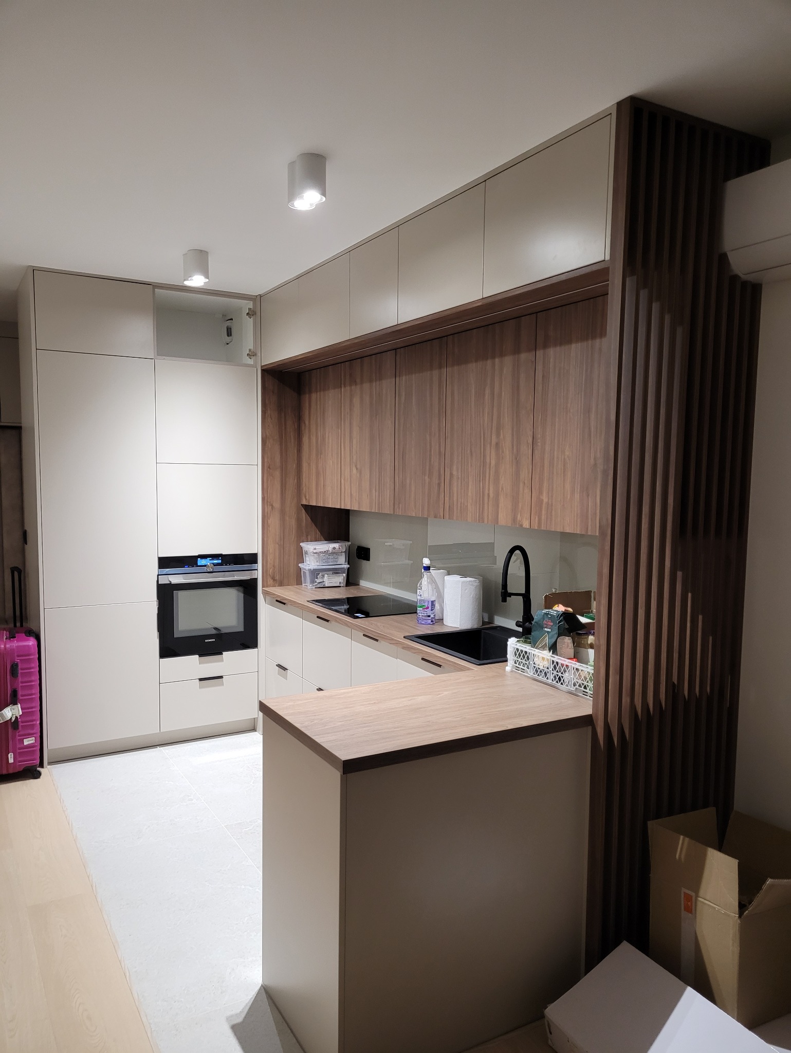 Nowoczesna kuchnia na wymiar z wyspą, fronty w odcieniach beżu i brązu, czarny zlew i armatura. Widoczny fragment ażurowej ścianki działowej.