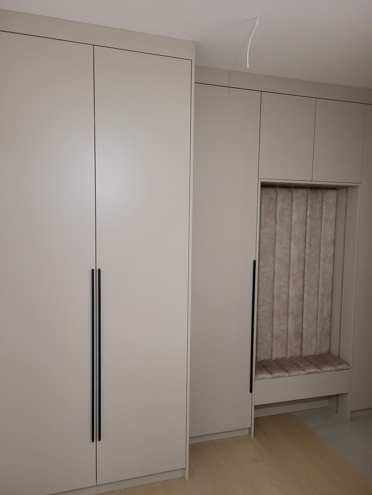 Jasnoszara szafa wnękowa z czarnymi uchwytami i wbudowanym siedziskiem z tapicerowanym oparciem. Minimalistyczny design, gładkie fronty, drewniana podłoga.