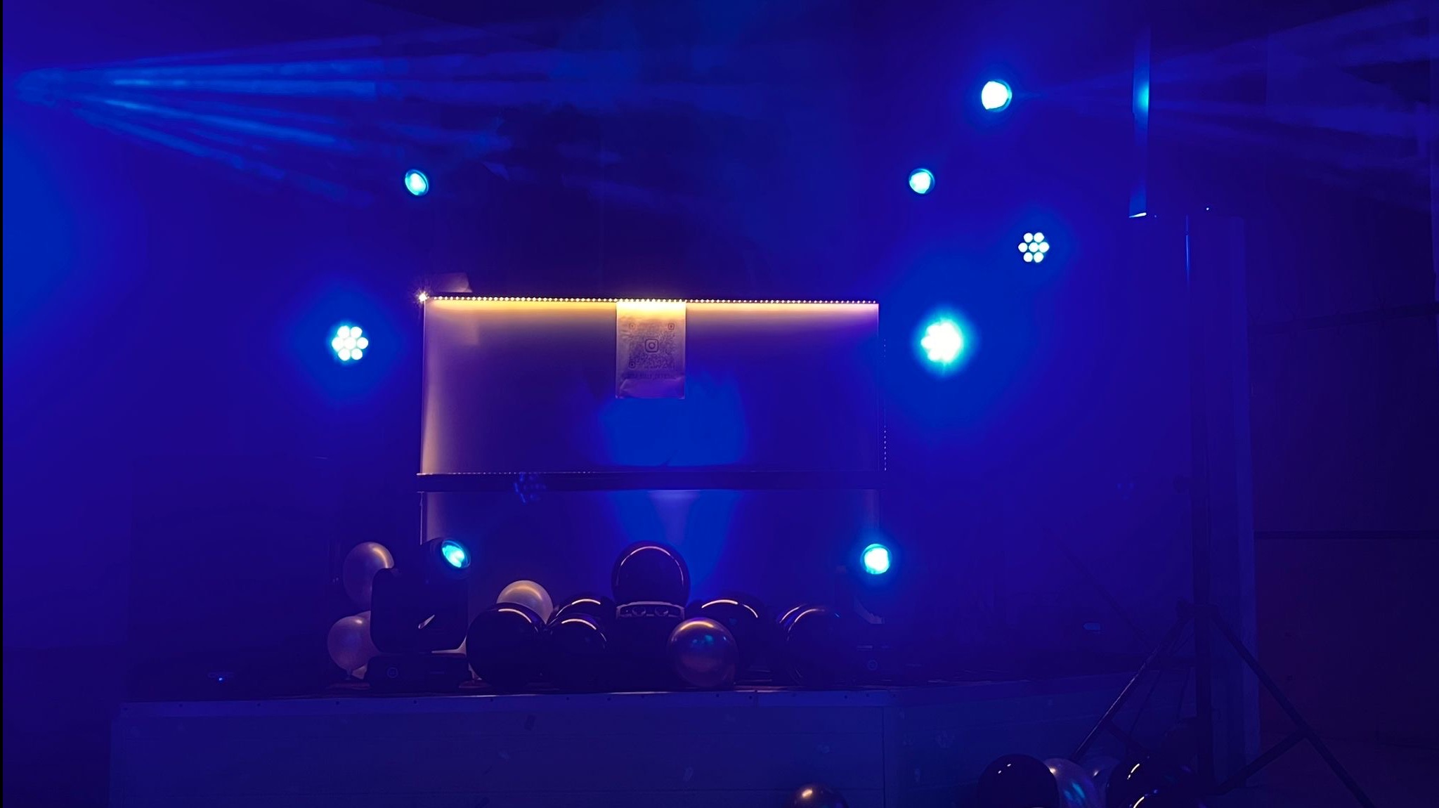 Scena w niebieskim oświetleniu z balonami, ekranem z kodem QR i reflektorami. Przygotowania do wydarzenia, widok z dołu.