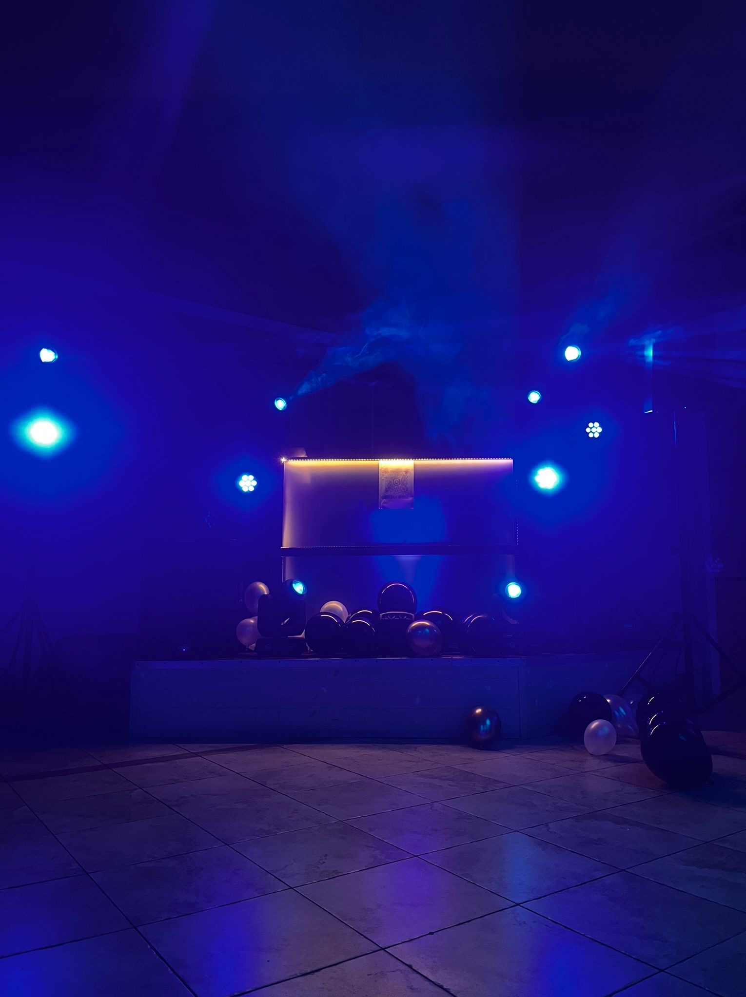 Scena oświetlona niebieskim światłem z dekoracjami z balonów, przygotowana na występ lub event. Widoczne reflektory i podświetlony panel.