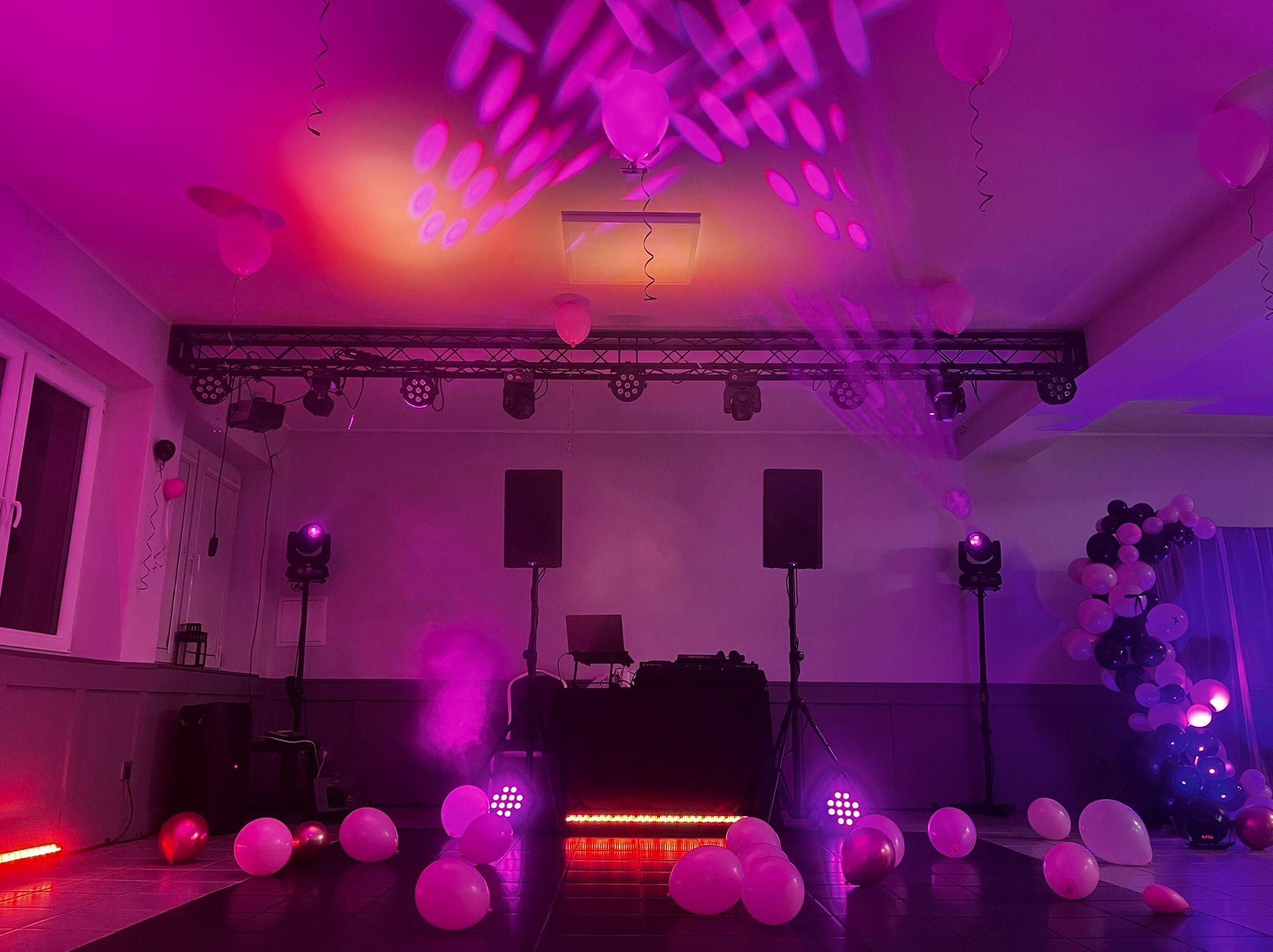 Oświetlona sala z DJ-ką, nagłośnieniem i dekoracjami z balonów w odcieniach różu i fioletu. Scena gotowa na imprezę, widoczne reflektory i sprzęt muzyczny.