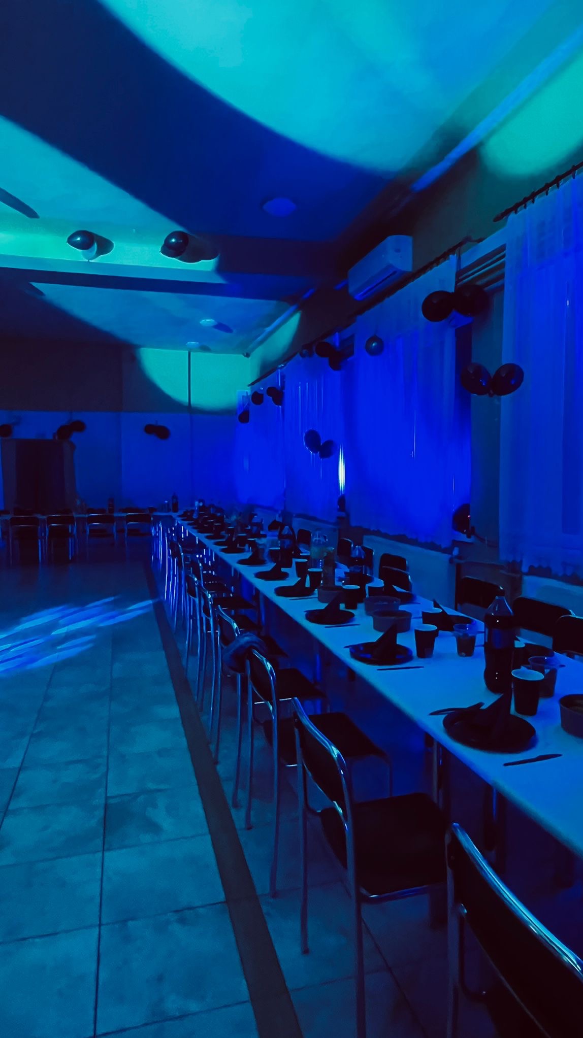 Uroczysta sala w niebieskim oświetleniu, stoły nakryte na czarno, dekoracje z balonów. Przygotowania do imprezy, widok z perspektywy gościa.