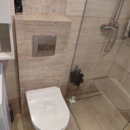 DUŻY MAŁY REMONT Damian Łoniewski - Wykończona łazienka z beżowymi płytkami, białą toaletą i szklaną kabiną prysznicową. Widoczny fragment szafki i dozownik mydła. Ujęcie z góry.