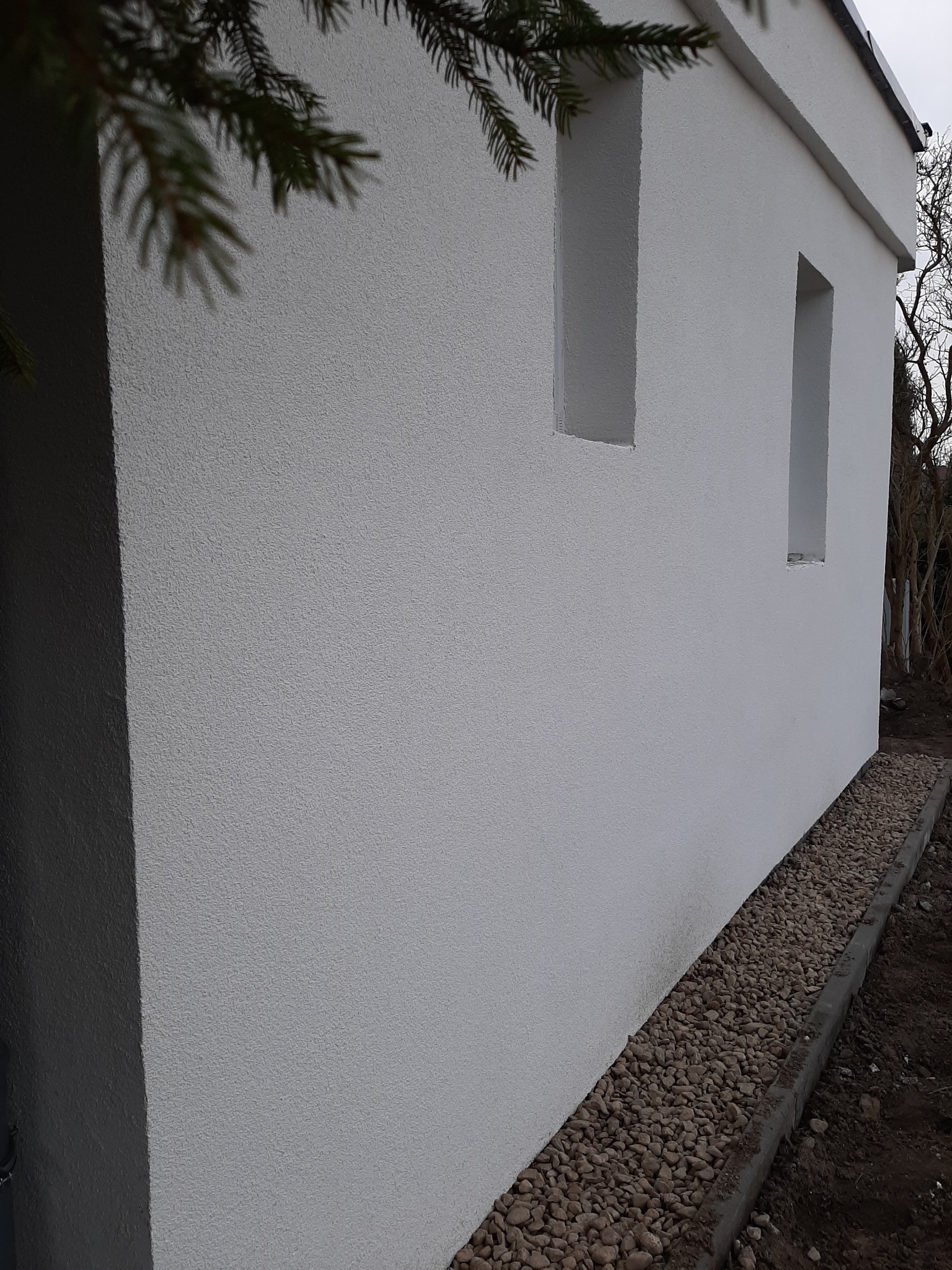 Biała elewacja budynku z fakturą baranka, dwa małe okna wnękowe, obok ścieżka z kamieni. Ujęcie z dołu, widoczne gałęzie drzewa iglastego.