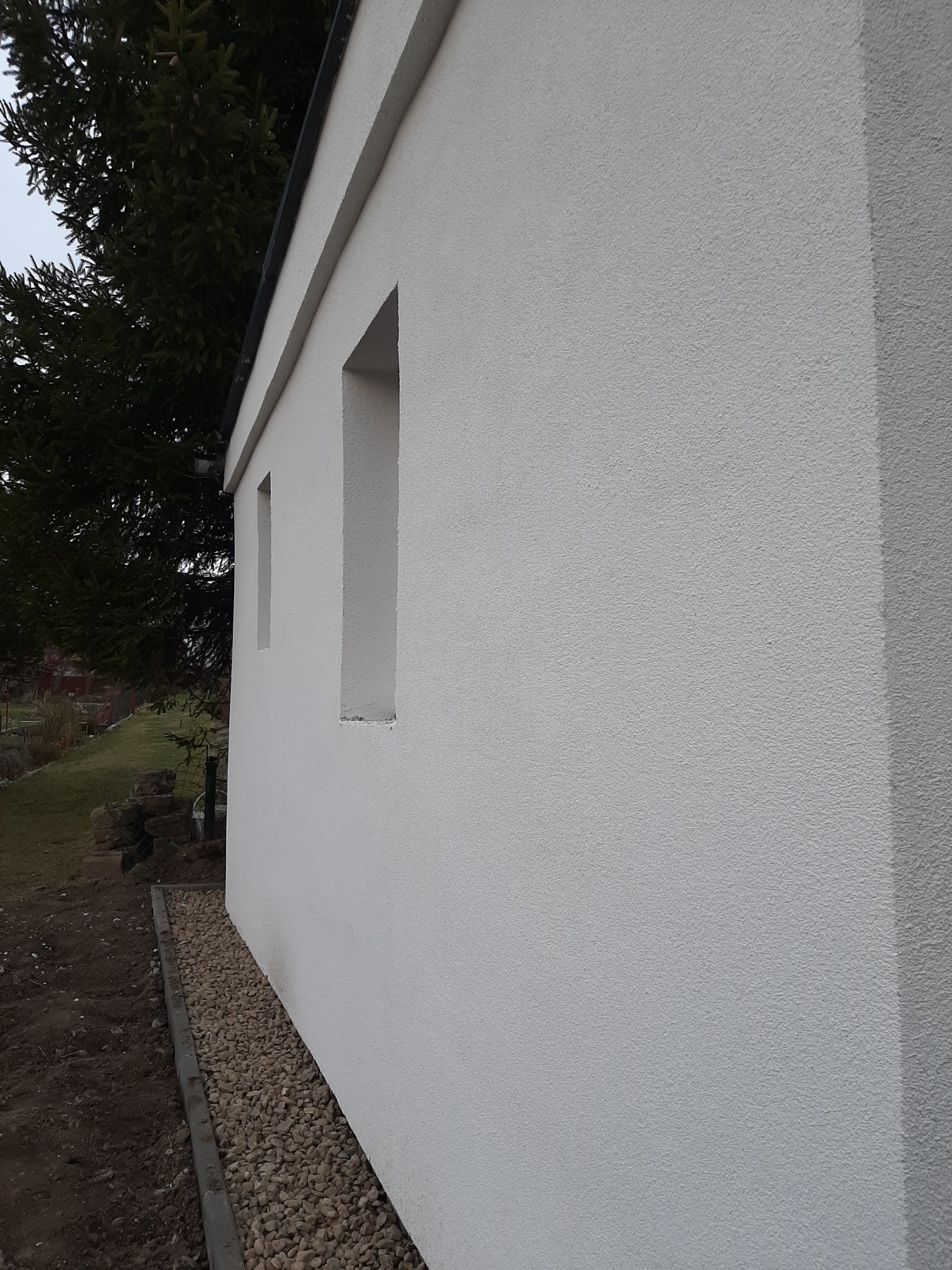 Biała elewacja budynku z małymi oknami, widok z boku. Obok budynku widoczny pas żwiru i fragment zieleni. Tekstura ściany chropowata.