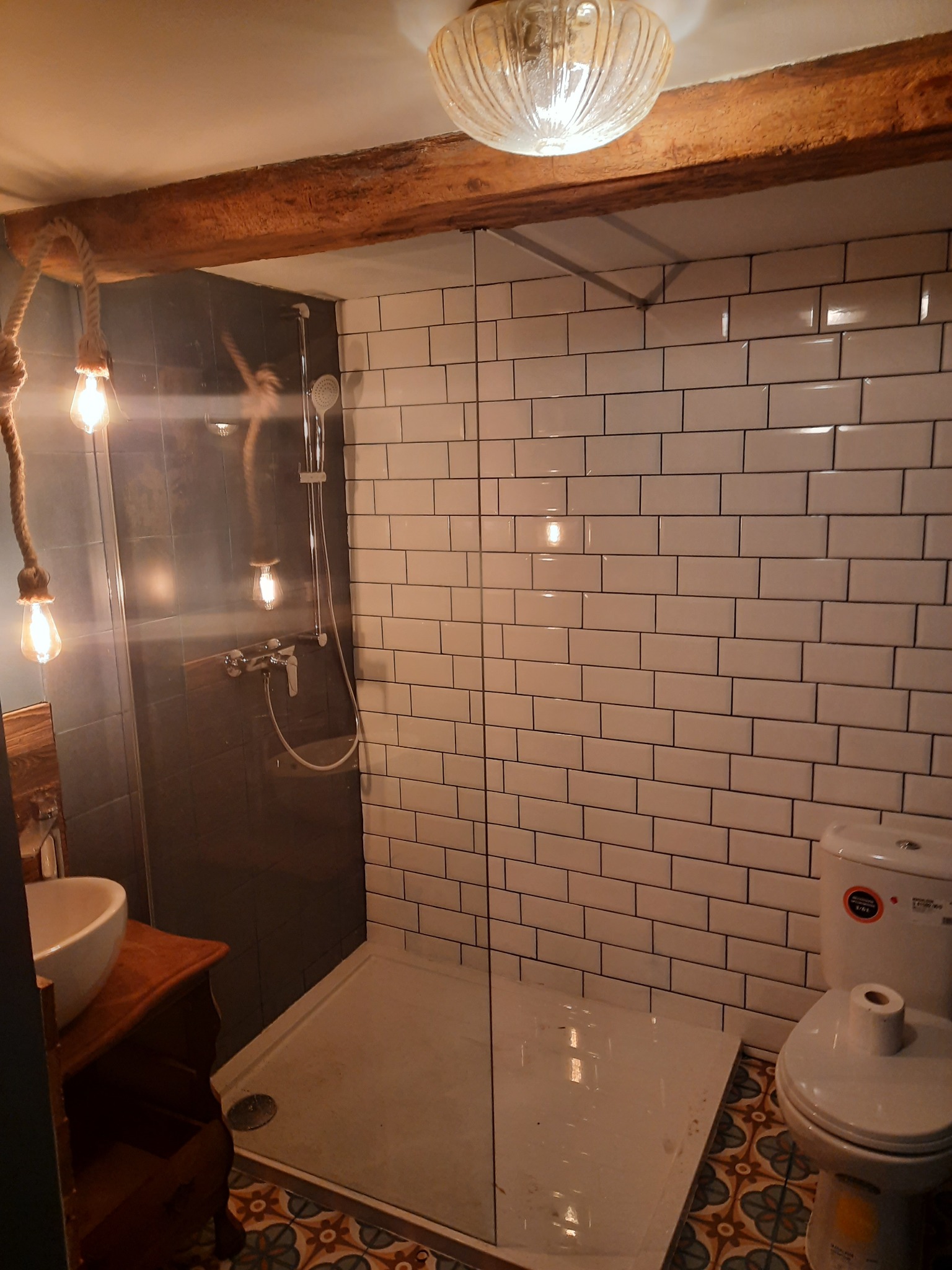 Wykończona łazienka w stylu retro: białe kafelki z czarną fugą, kabina prysznicowa, umywalka na drewnianej komodzie, dekoracyjne oświetlenie i wzorzysta podłoga.