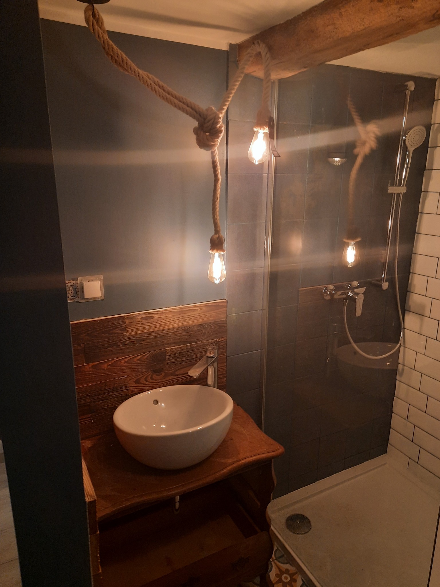 Rustykalna łazienka z drewnianą umywalką, szarą ścianą i kabiną prysznicową. Lampa na sznurze dodaje unikalnego charakteru. Widoczne płytki i armatura.