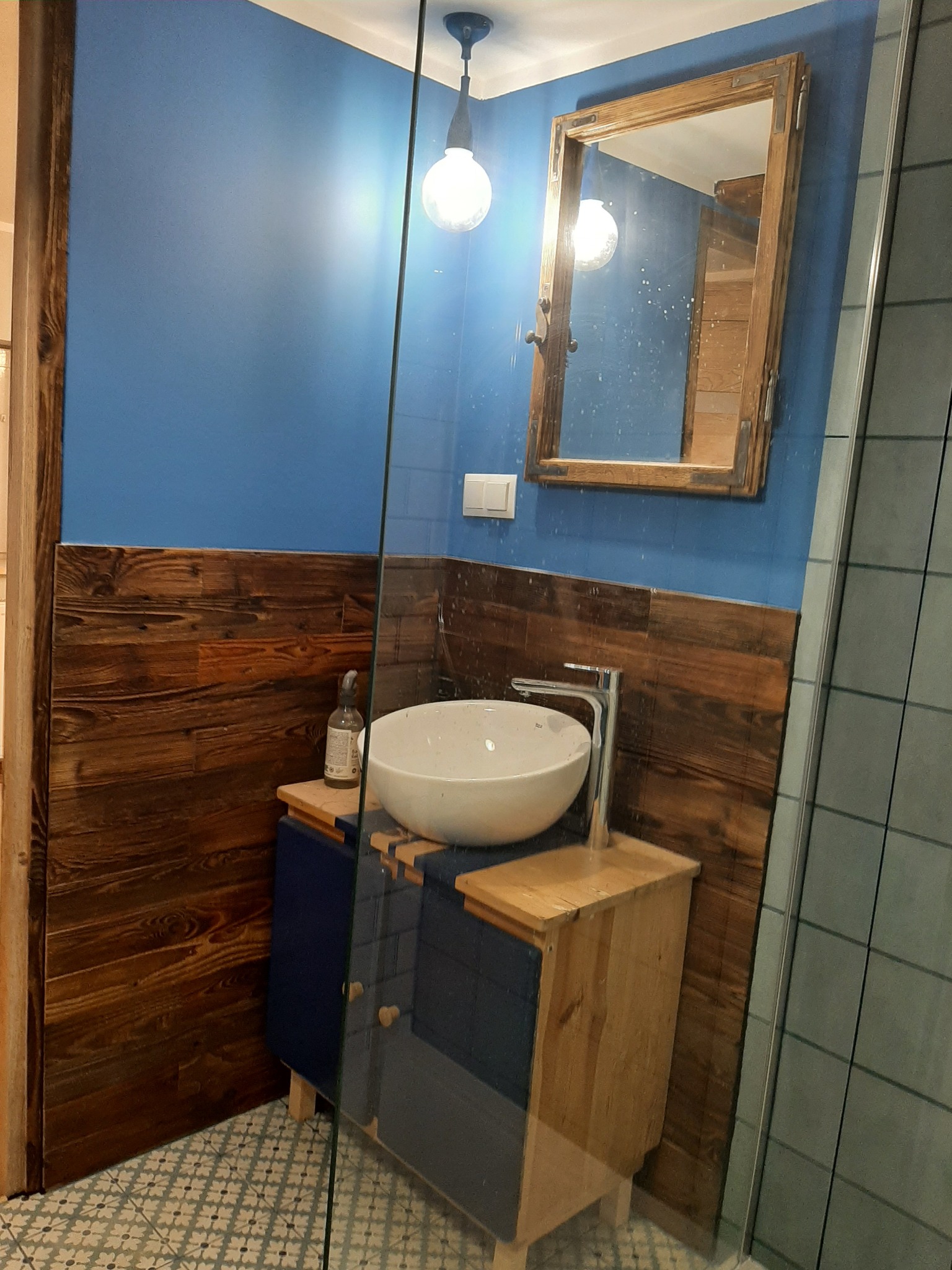 Niebieska ściana nad drewnianą boazerią w łazience z umywalką na szafce z drewnianymi i niebieskimi elementami, lustro w drewnianej ramie i szklana kabina prysznicowa.
