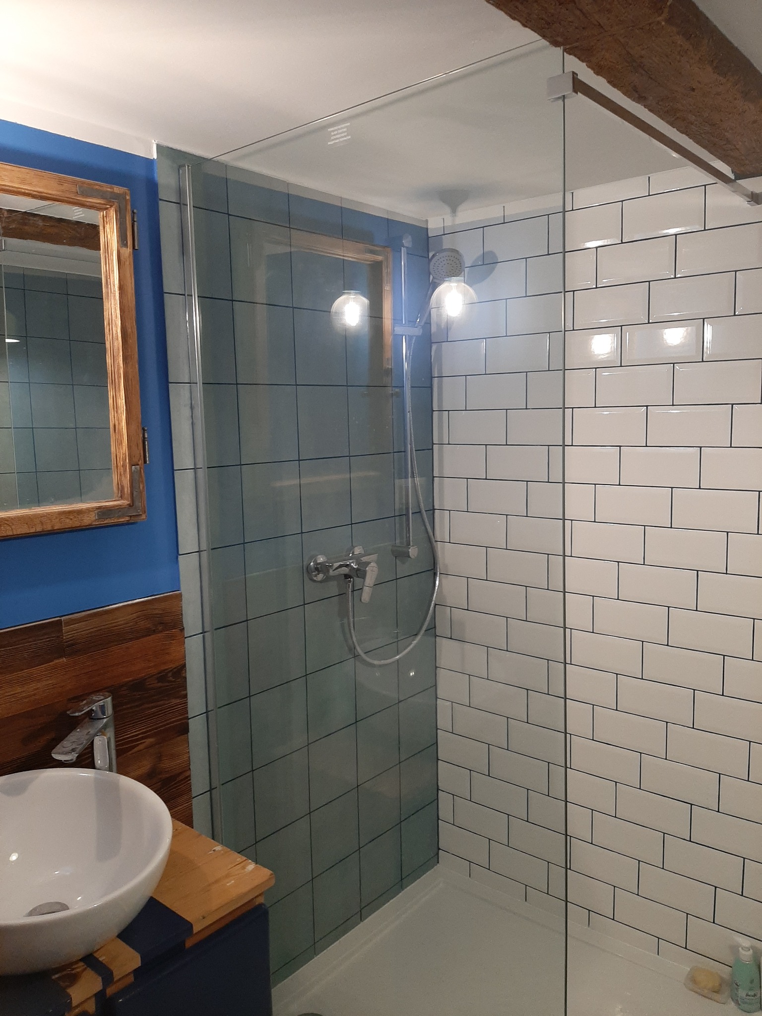 Wykończona łazienka z kabiną prysznicową oddzieloną szklaną ścianką, białe płytki imitujące cegłę, drewniane elementy i niebieska ściana z lustrem w drewnianej ramie.