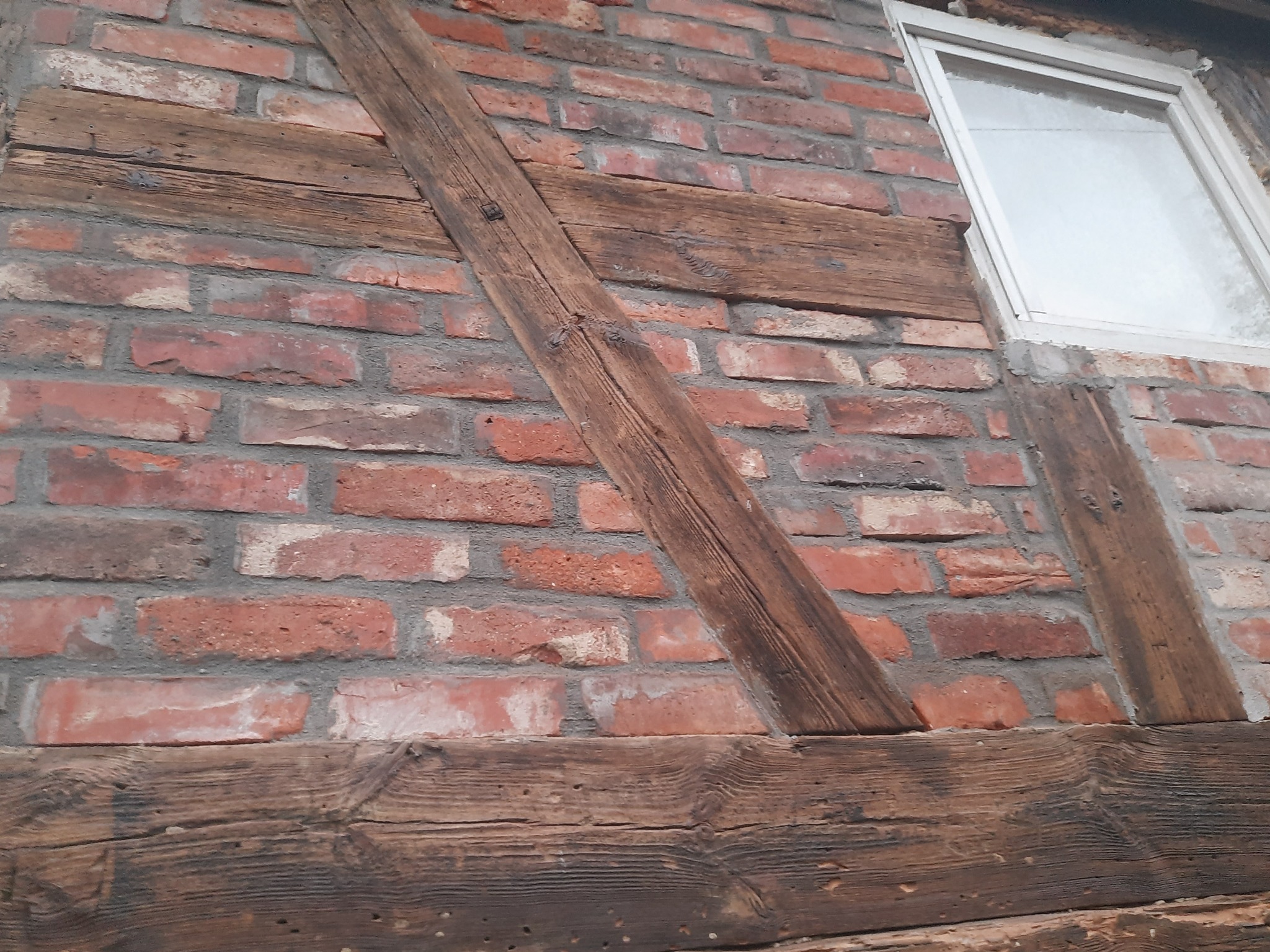 Ceglana ściana z drewnianymi elementami konstrukcyjnymi i oknem. Widoczne detale faktury cegieł i drewna, tworzące rustykalny wygląd elewacji.