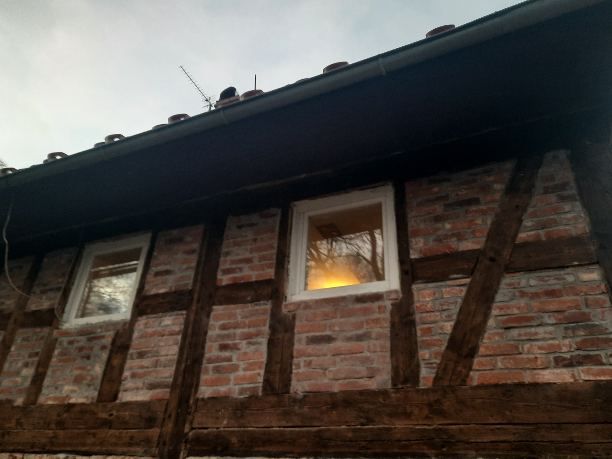 Fragment elewacji domu z muru pruskiego z oknami, widok z dołu na dach i antenę. Ceglana ściana z drewnianymi belkami konstrukcyjnymi.
