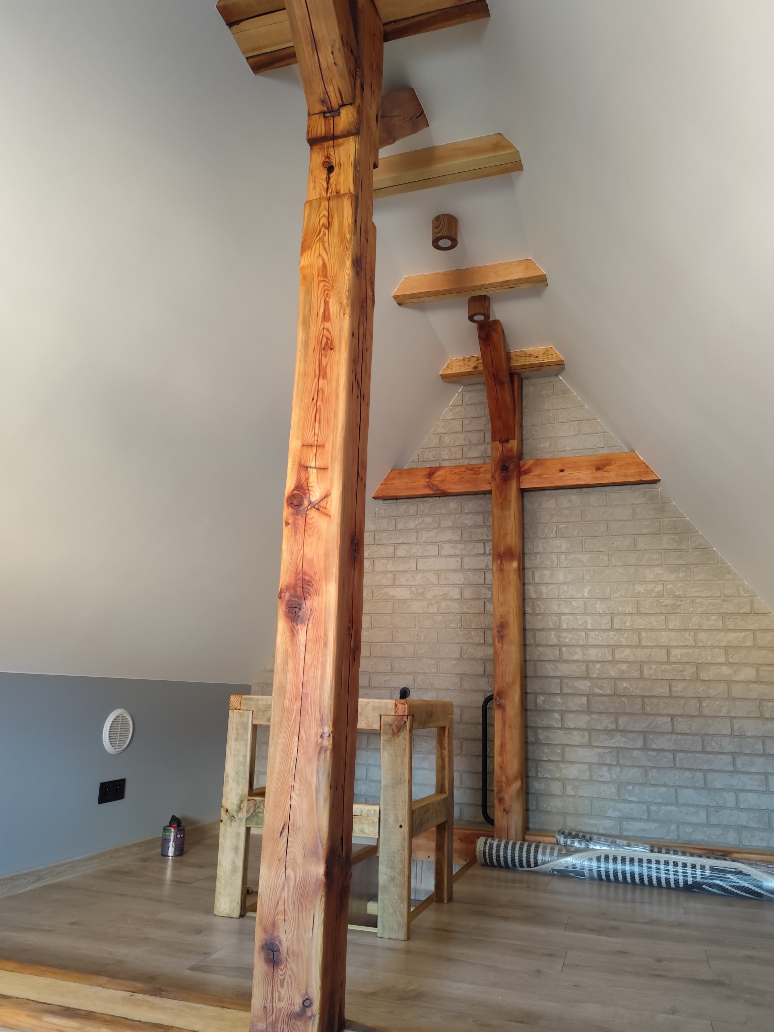 Drewniane belki konstrukcyjne w jasnym poddaszu z ceglaną ścianą i drewnianą podłogą. Widoczne elementy oświetlenia i materiały budowlane.