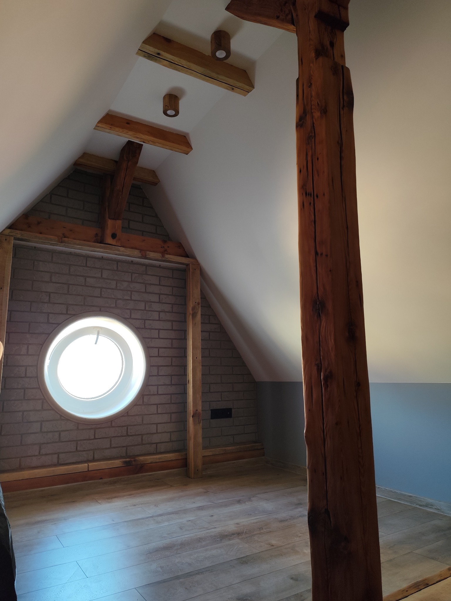 Adaptacja poddasza: okrągłe okno w ceglanej ścianie szczytowej, drewniane belki stropowe z oświetleniem i rustykalny słup na tle szarej ściany i drewnianej podłogi.