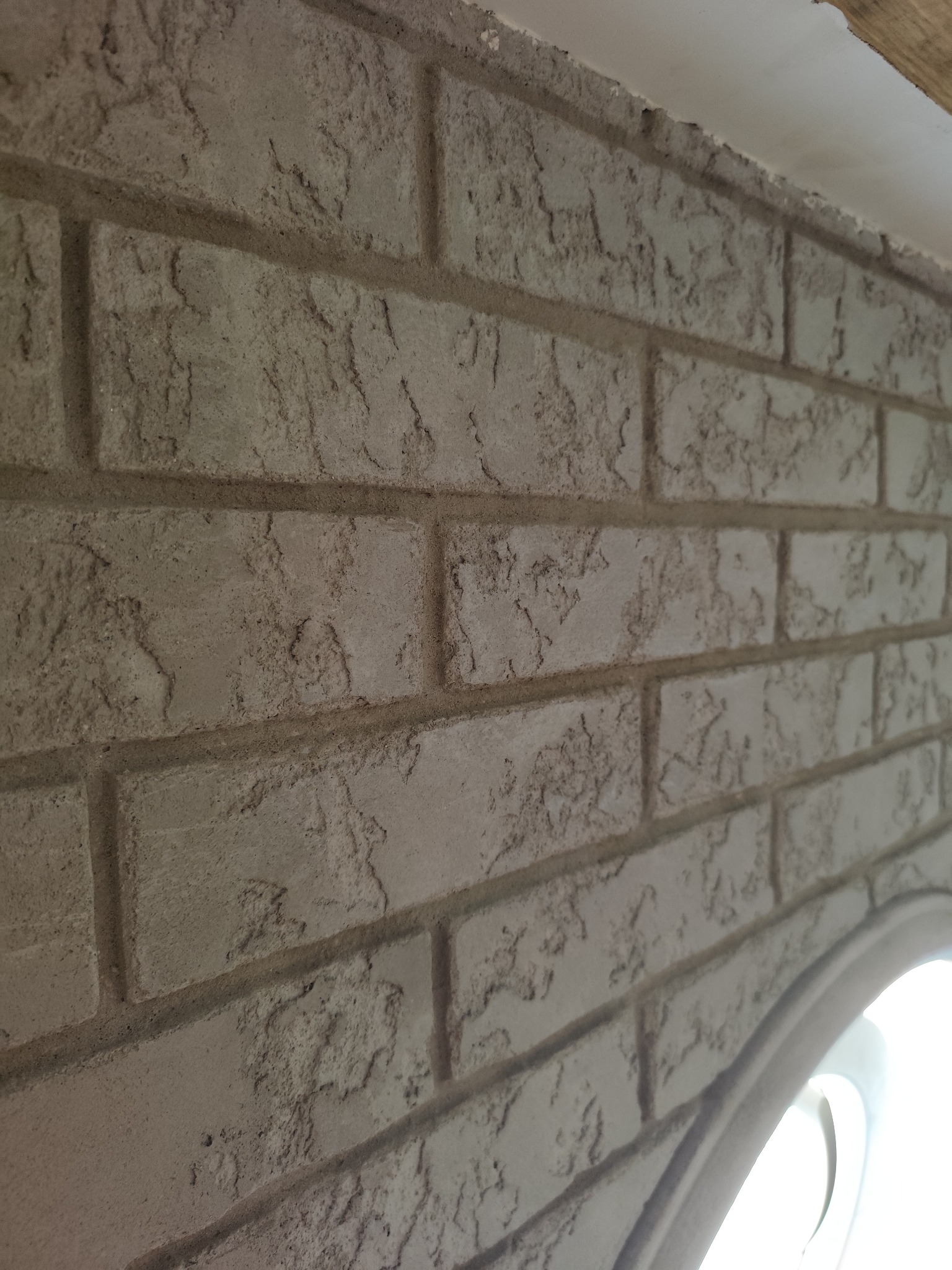 Dekoracyjna ściana z efektem cegły w odcieniach szarości, z wyraźną fakturą i fugami. Widoczny fragment okna z zaokrąglonym łukiem.