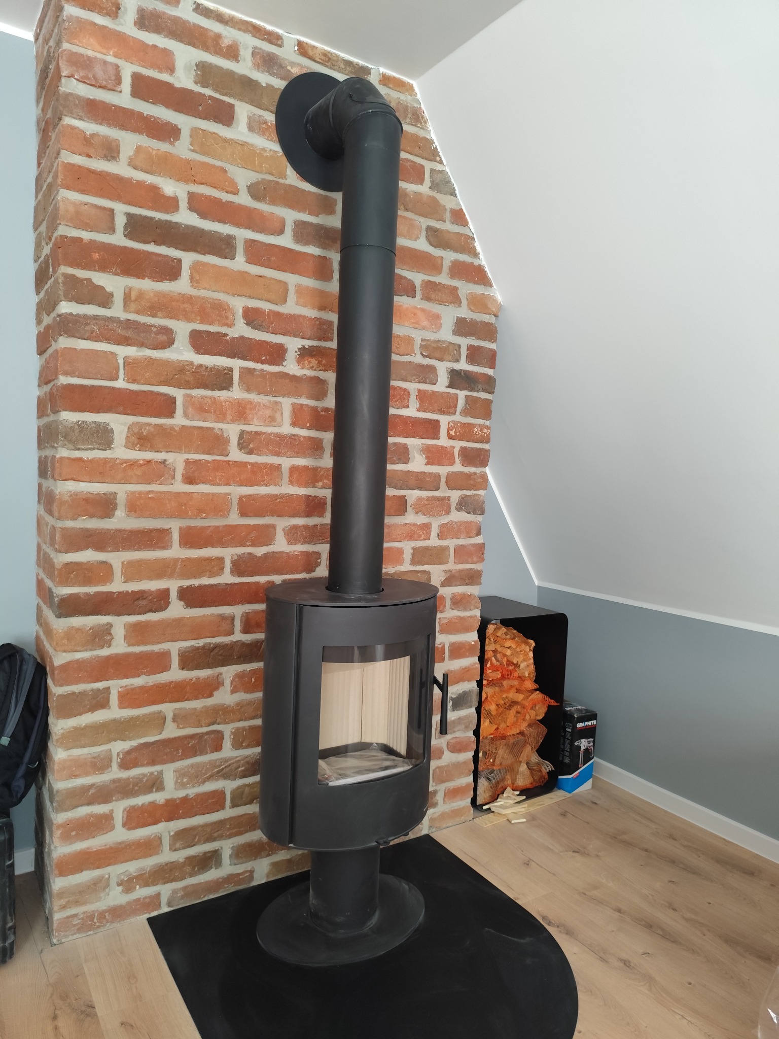 Ceglana ściana z kominem w salonie z drewnianą podłogą. Komin w kolorze czarnym. Dekoracja z drewnem obok kominka. Ściana w kolorze ceglastym.