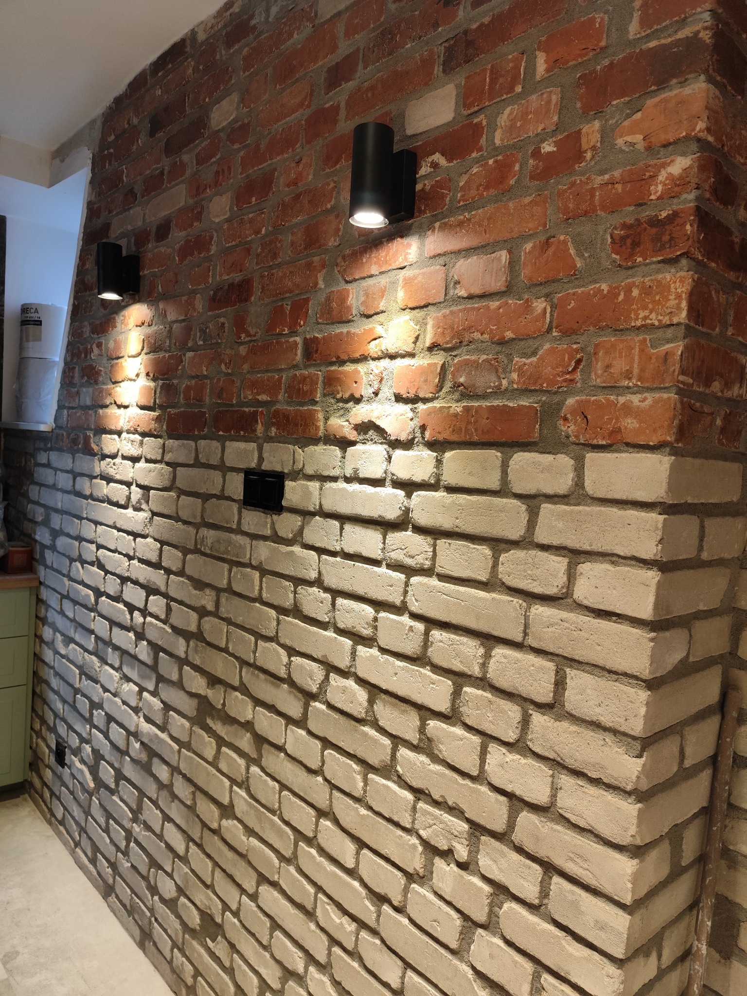 Ściana z cegły, częściowo pomalowana na biało, oświetlona dwoma czarnymi lampami ściennymi. Widoczny fragment instalacji elektrycznej. Dekoracyjne wykończenie wnętrza.
