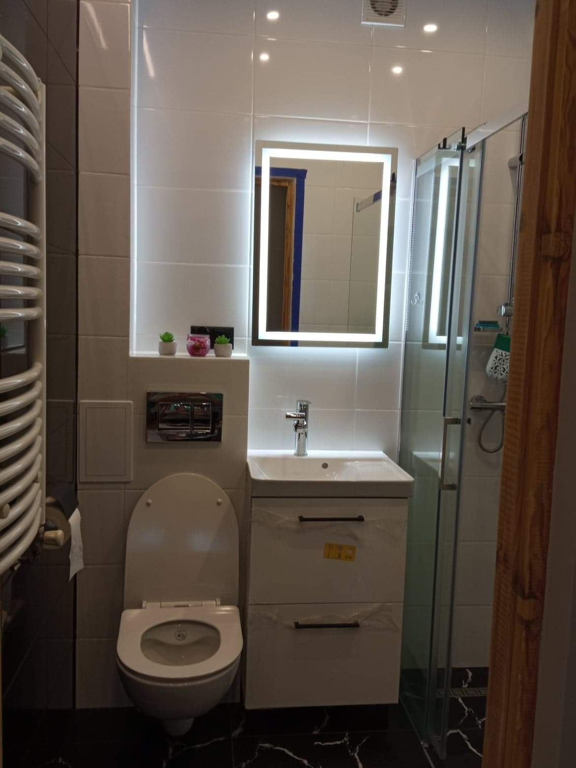 Nowoczesna łazienka z podświetlanym lustrem, białą armaturą i czarną podłogą. Umywalka z szafką w folii ochronnej. Widoczna kabina prysznicowa i grzejnik drabinkowy.