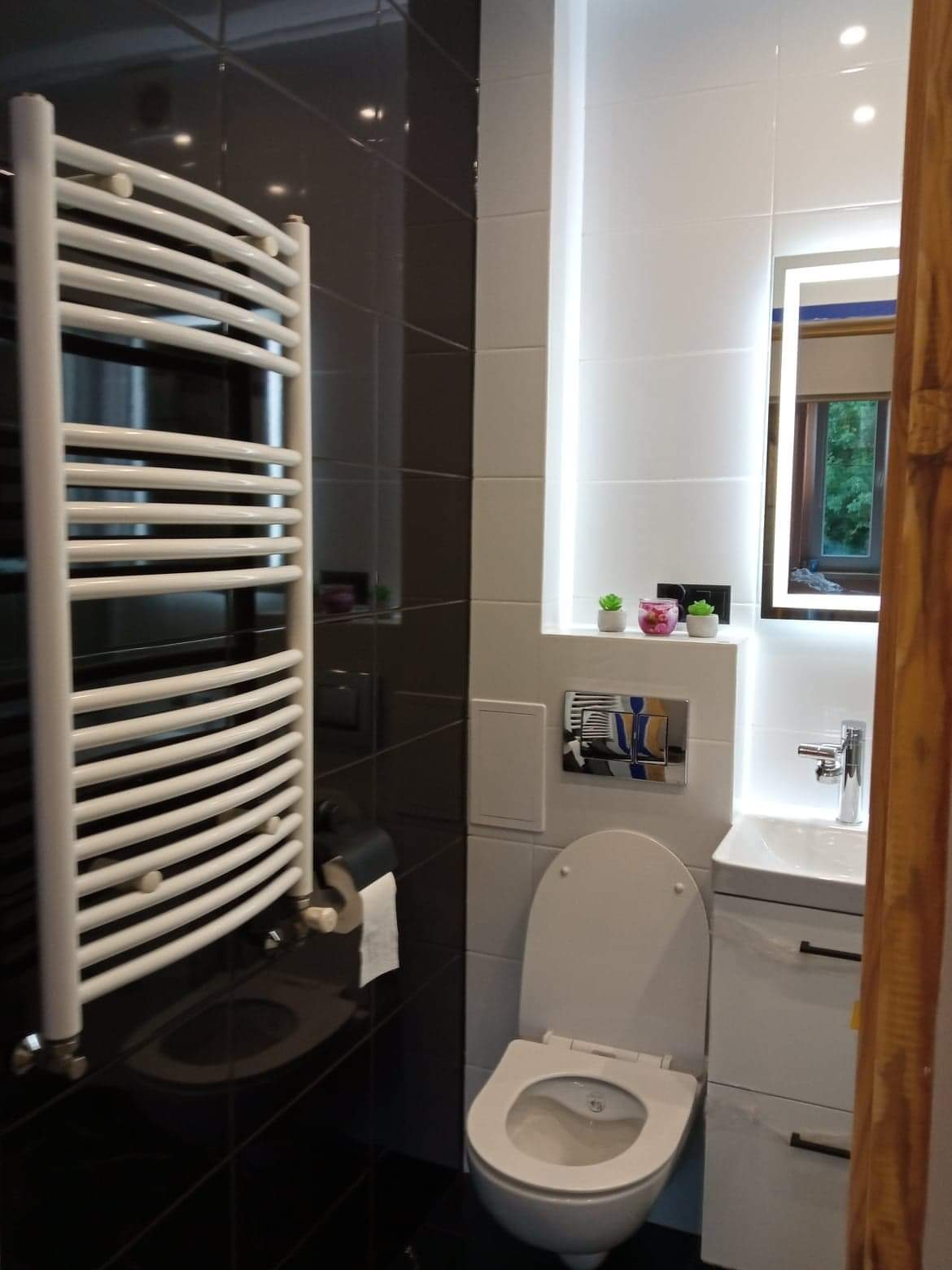 Nowoczesna łazienka z czarno-białymi płytkami, podświetlanym lustrem i grzejnikiem drabinkowym. Minimalistyczny design, czysta armatura, małe rośliny dekoracyjne.