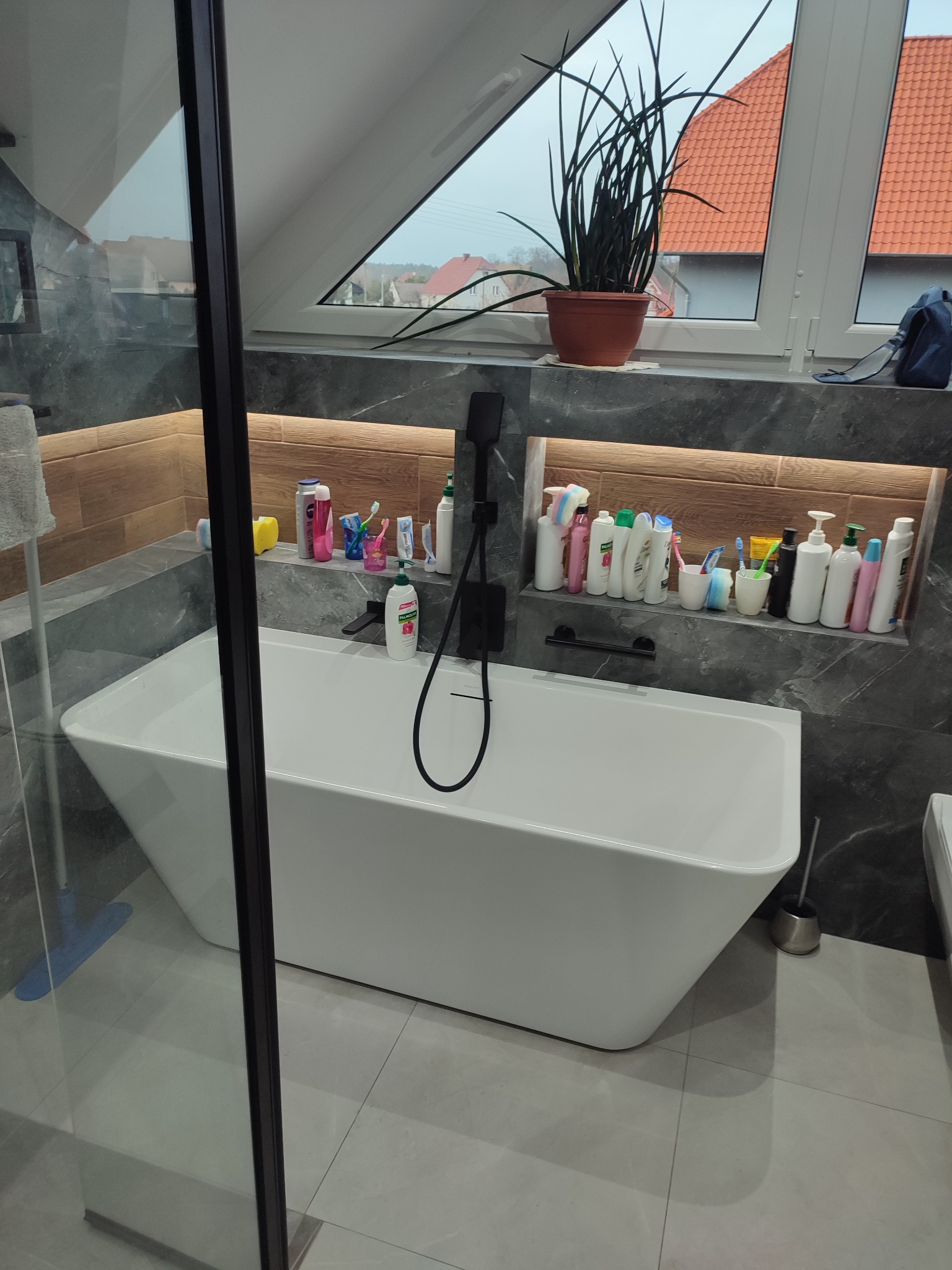 Nowoczesna łazienka na poddaszu z wolnostojącą białą wanną, szarą okładziną imitującą marmur i drewnianą wnęką z kosmetykami. Widok przez szklaną kabinę.