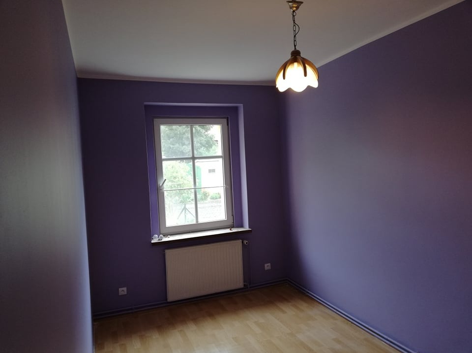 Pomalowane na fioletowo wnętrze pokoju z drewnianą podłogą, białym oknem i grzejnikiem. Na suficie wisi lampa w stylu retro.