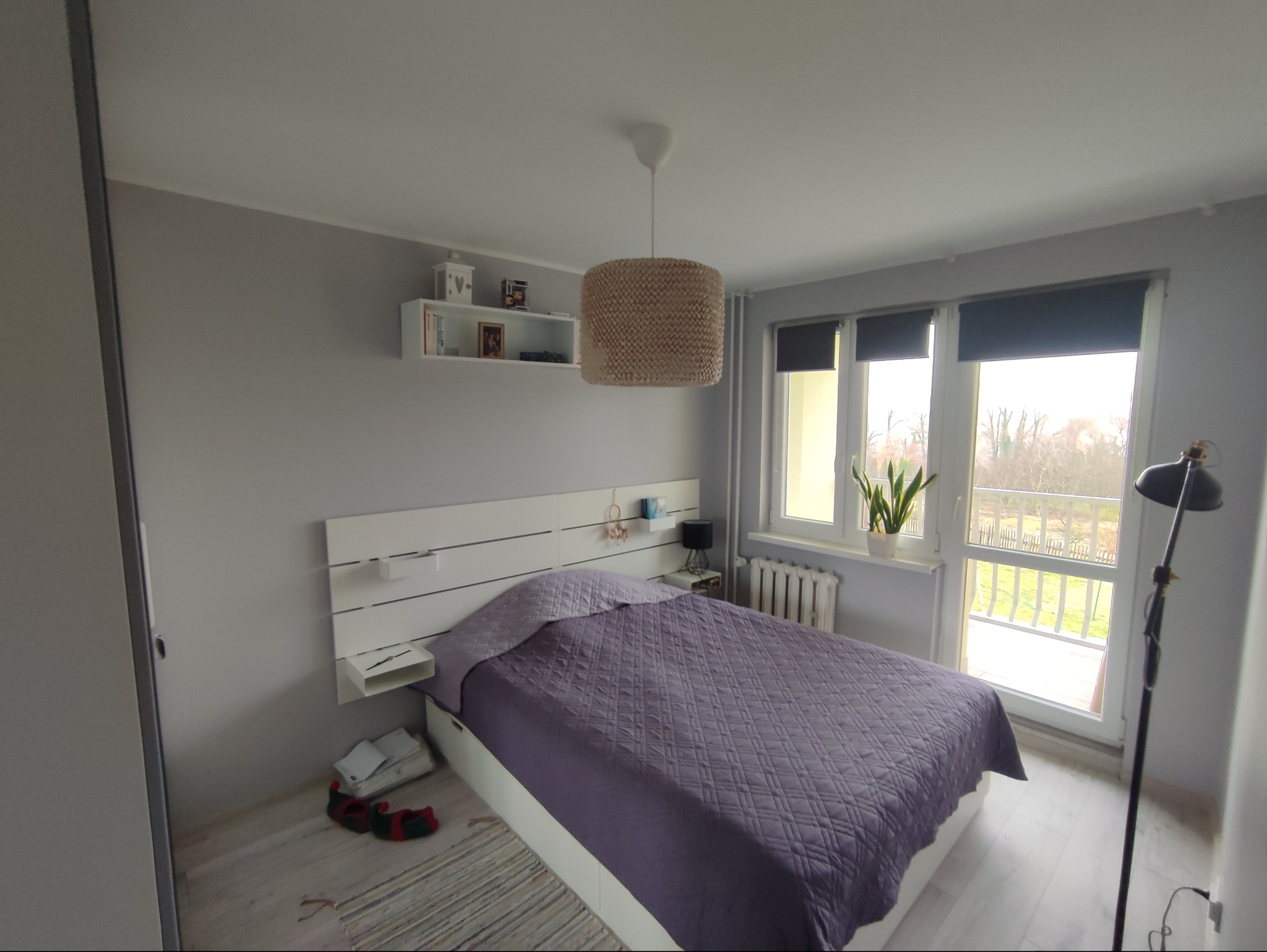 Jasna sypialnia z fioletową narzutą na łóżku, białym zagłówkiem z półkami i lampą z abażurem. Widok na balkon przez okno z roletami. Minimalistyczny styl.