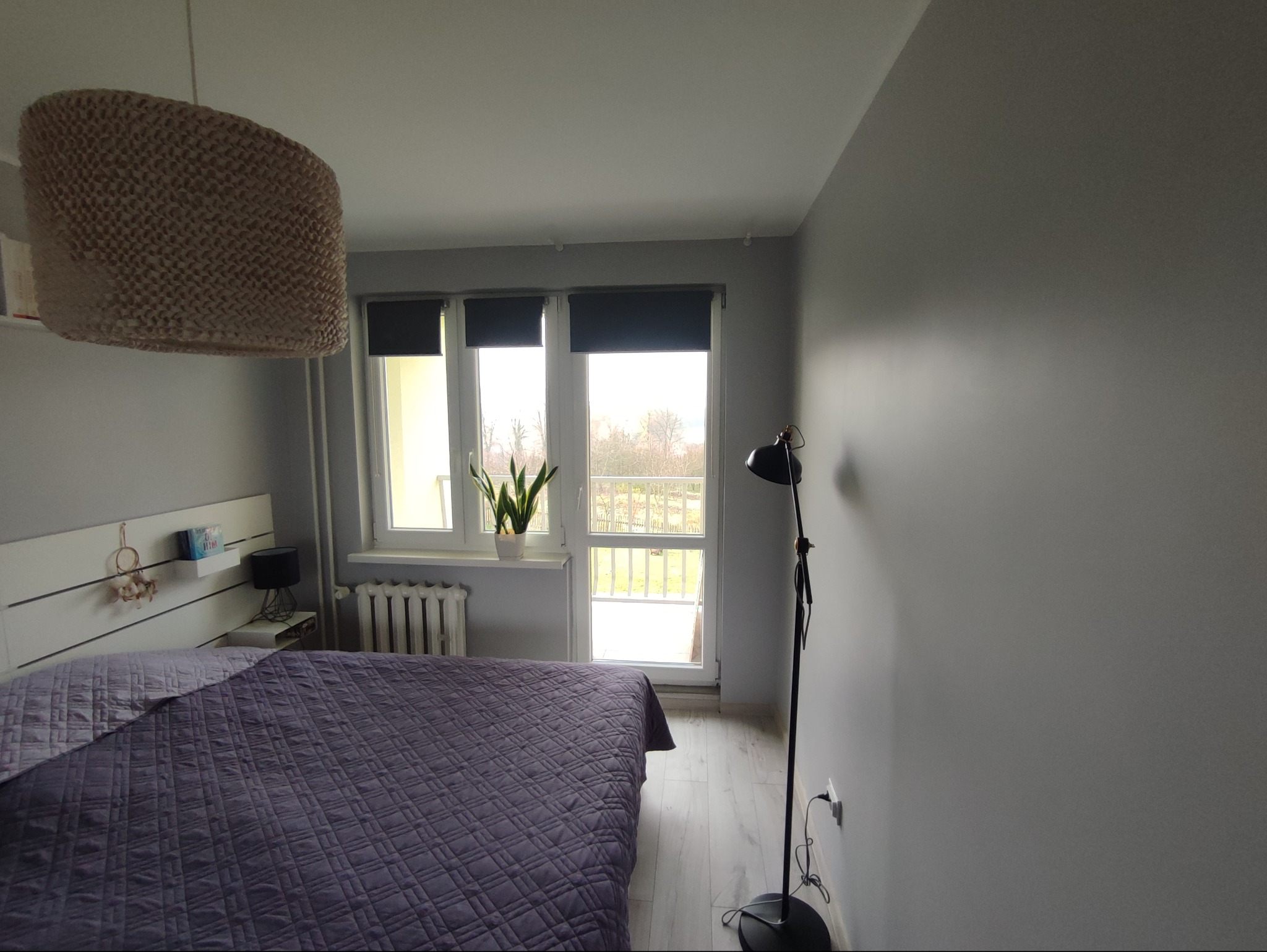 Jasna sypialnia z fioletową narzutą na łóżku, lampą podłogową i widokiem na balkon przez okno z roletami. Minimalistyczny wystrój, szare ściany.