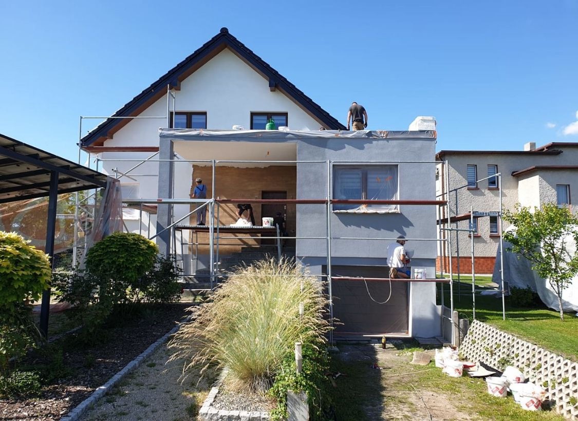 Dom jednorodzinny w trakcie malowania elewacji na szaro, z widocznymi rusztowaniami i pracownikami. Część ściany jeszcze w surowym stanie. Słoneczny dzień.