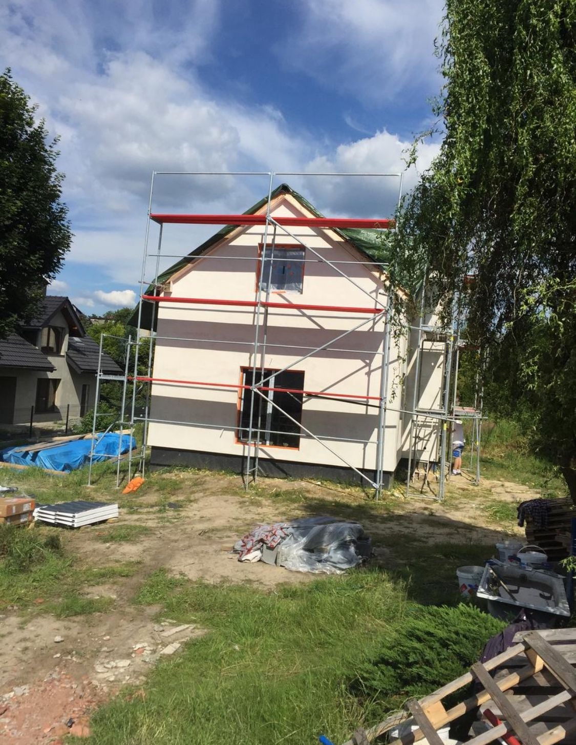 Dom jednorodzinny w trakcie prac elewacyjnych, z widocznym rusztowaniem i przygotowanymi materiałami budowlanymi. Świeżo otynkowana fasada w jasnym odcieniu.