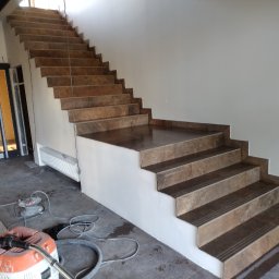 Mykhailo Hnatiuk - Wykończone schody z brązowych płytek z antypoślizgowymi paskami, widoczne narzędzia pracy i odkurzacz przemysłowy. Jasne wnętrze w trakcie prac wykończeniowych.