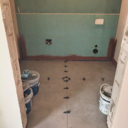 Mykhailo Hnatiuk - Przygotowanie do układania płytek w łazience: widoczne poziomowanie podłoża z użyciem krzyżyków dystansowych, ściany z płyt kartonowo-gipsowych i pojemniki z materiałami.