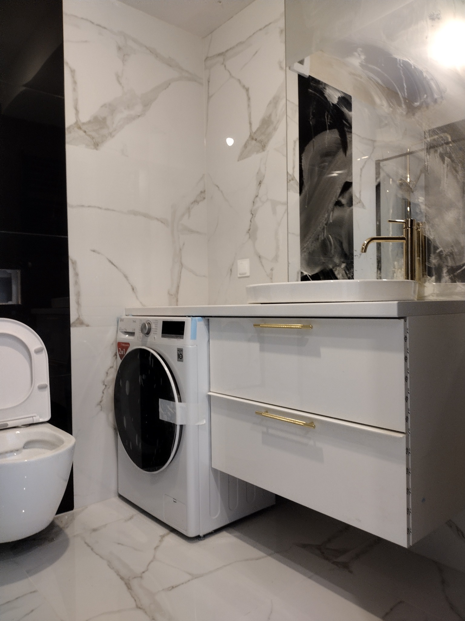 Nowoczesna łazienka z marmurowymi płytkami, białą szafką z umywalką i złotą armaturą. Widoczna pralka i toaleta. Minimalistyczny design z akcentami luksusu.