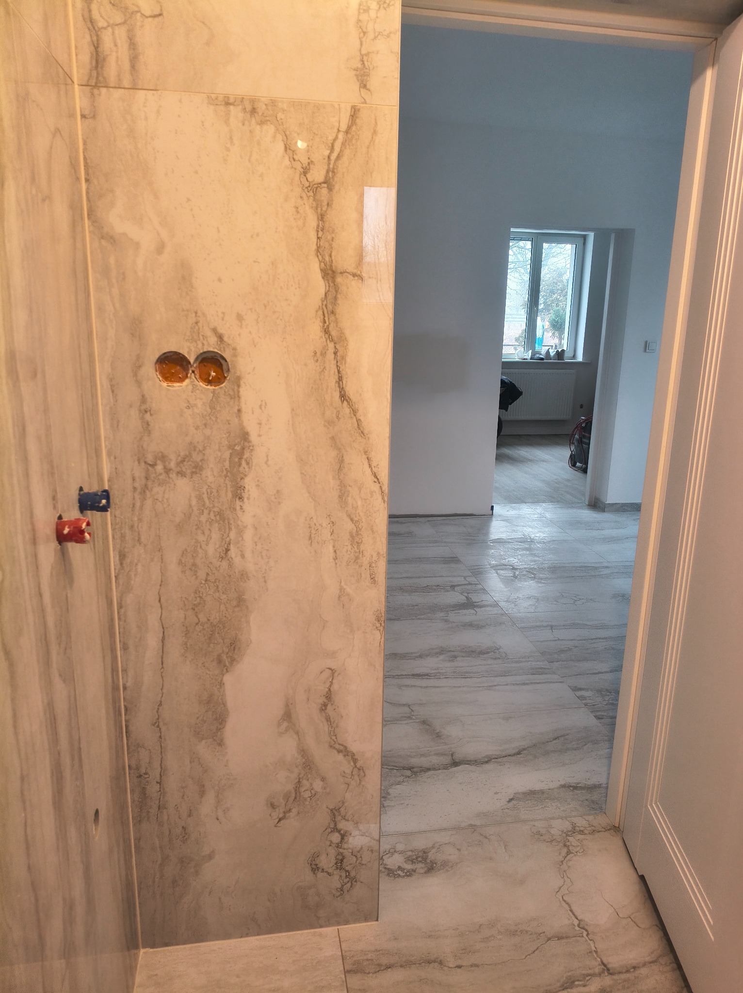 Wykończona łazienka z marmurowymi płytkami, widoczne przyłącza wodne i podłoga. W tle widok na pokój z oknem. Minimalistyczny design i jasna kolorystyka.