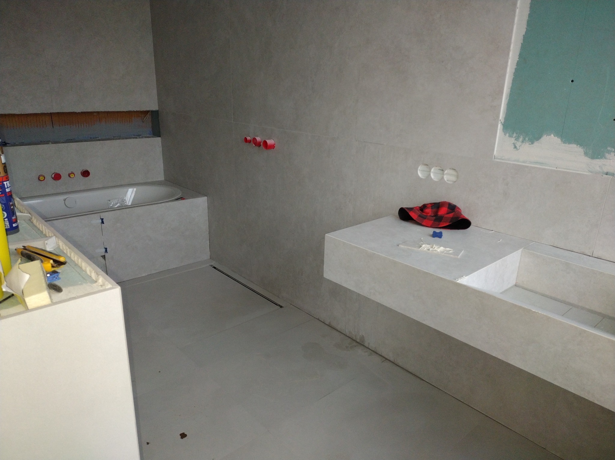 Wnętrze łazienki w trakcie wykańczania: szare płytki, zabudowana wanna, umywalka z blatem, widoczne narzędzia i akcesoria. Czerwona czapka na blacie dodaje koloru.
