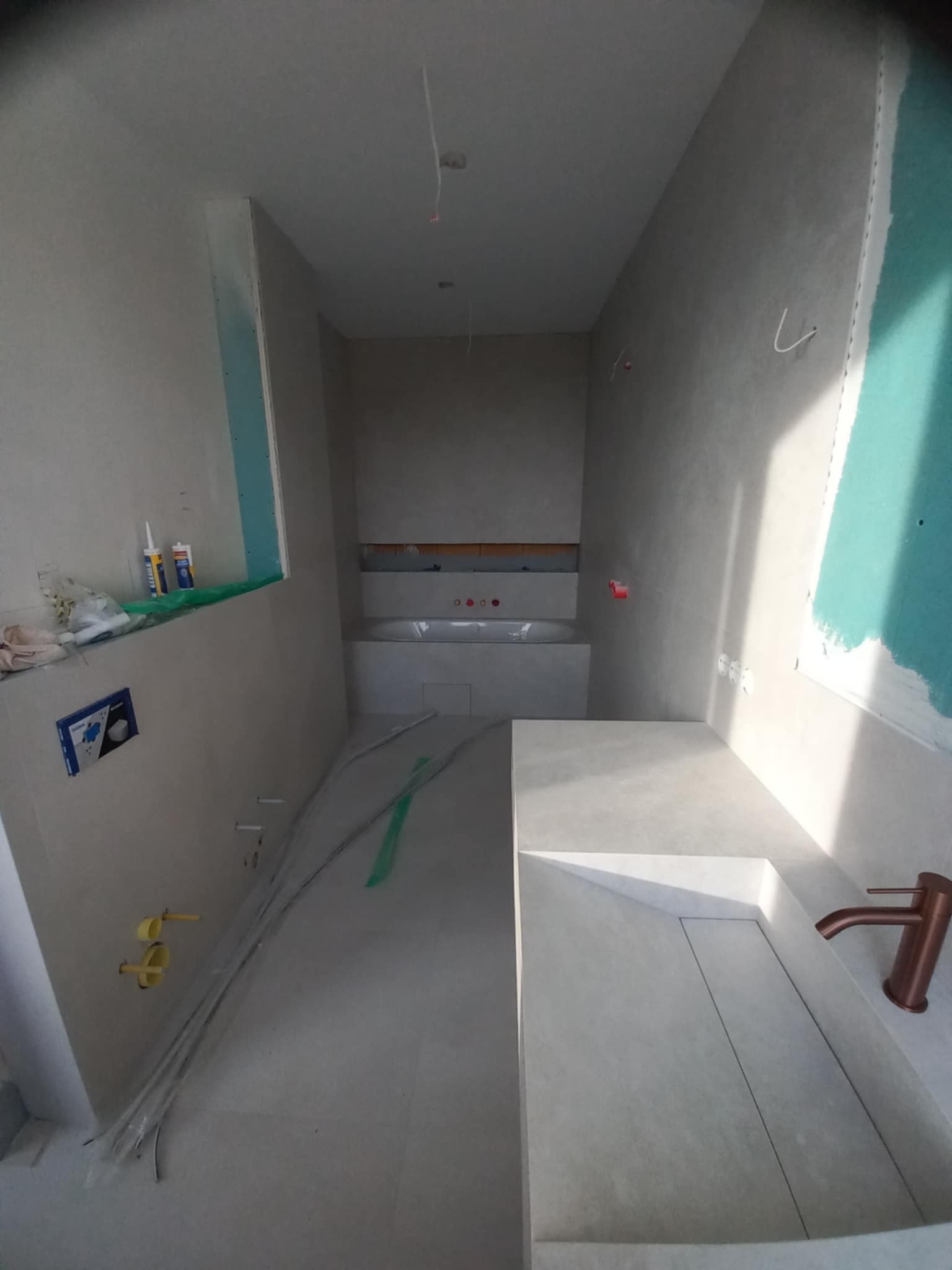 Wnętrze łazienki w trakcie prac wykończeniowych: widoczne płyty gipsowo-kartonowe, instalacja wodno-kanalizacyjna i umywalka z baterią w kolorze miedzi.