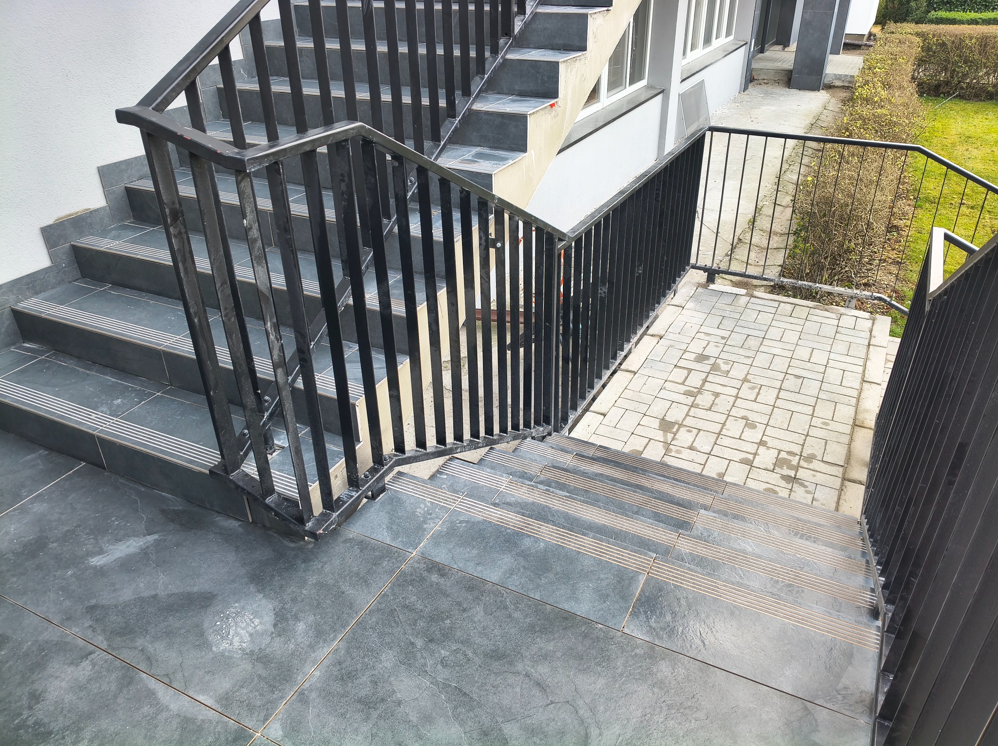 Zewnętrzne schody wyłożone szarą, matową płytką z antypoślizgowymi paskami. Metalowa, czarna balustrada. Widok z góry na fragment budynku i zieleni.