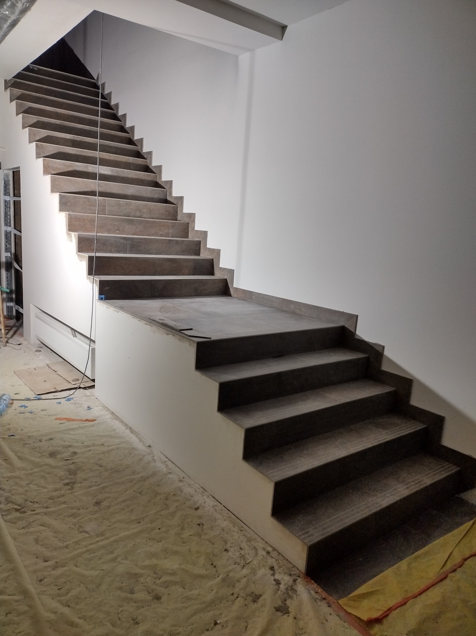 Wykończone schody z szarymi płytkami, białe ściany i podstopnice. Podłoga zabezpieczona folią malarską. Widoczne elementy instalacji i narzędzia.