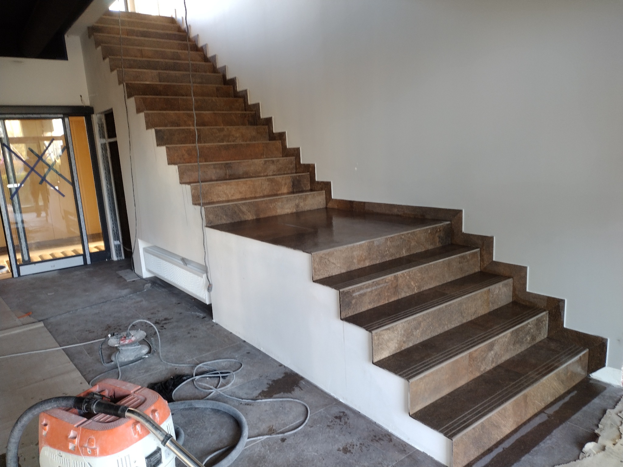 Wykończone schody z brązowych płytek z antypoślizgowymi paskami, widoczne narzędzia pracy i odkurzacz przemysłowy. Jasne wnętrze w trakcie prac wykończeniowych.