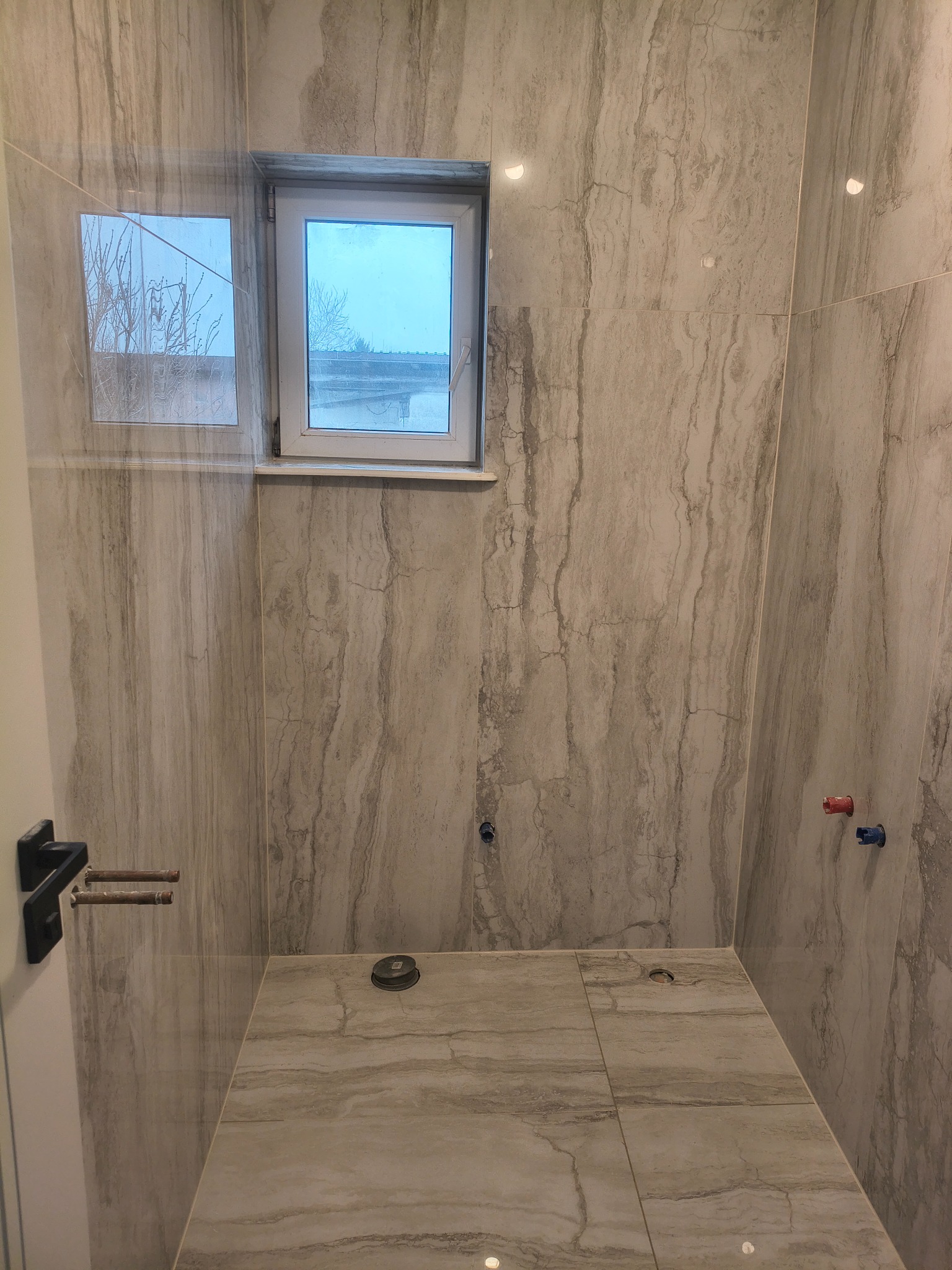 Wykończona łazienka z marmurowymi płytkami, widoczne okno i przygotowane instalacje. Minimalistyczny design z elementami w kolorze szarym i białym.