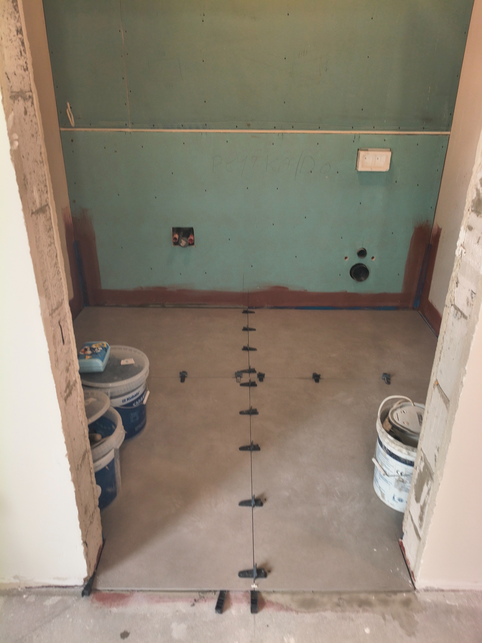 Przygotowanie do układania płytek w łazience: widoczne poziomowanie podłoża z użyciem krzyżyków dystansowych, ściany z płyt kartonowo-gipsowych i pojemniki z materiałami.