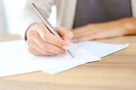 Dłoń kobiety piszącej srebrnym długopisem na białej kartce papieru leżącej na jasnym drewnianym biurku. Ujęcie z bliska, skupione na dłoni i piśmie.